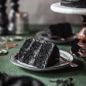 A slice of black velvet cake on a green table next to a cake stand, black linen, and candles.