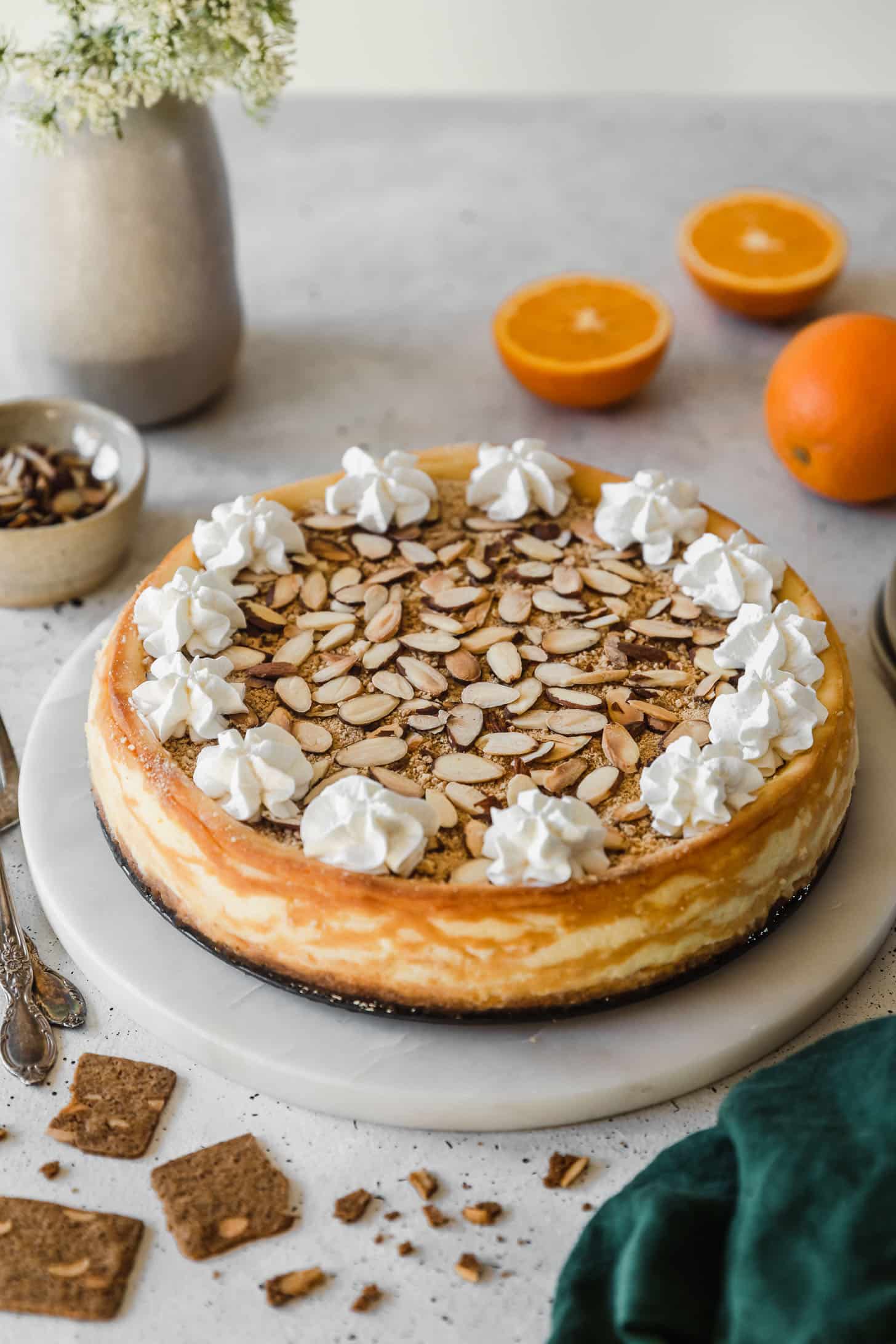 A cheesecake on a white table next to oranges, an emerald green linen, and a flower vase of white buds.