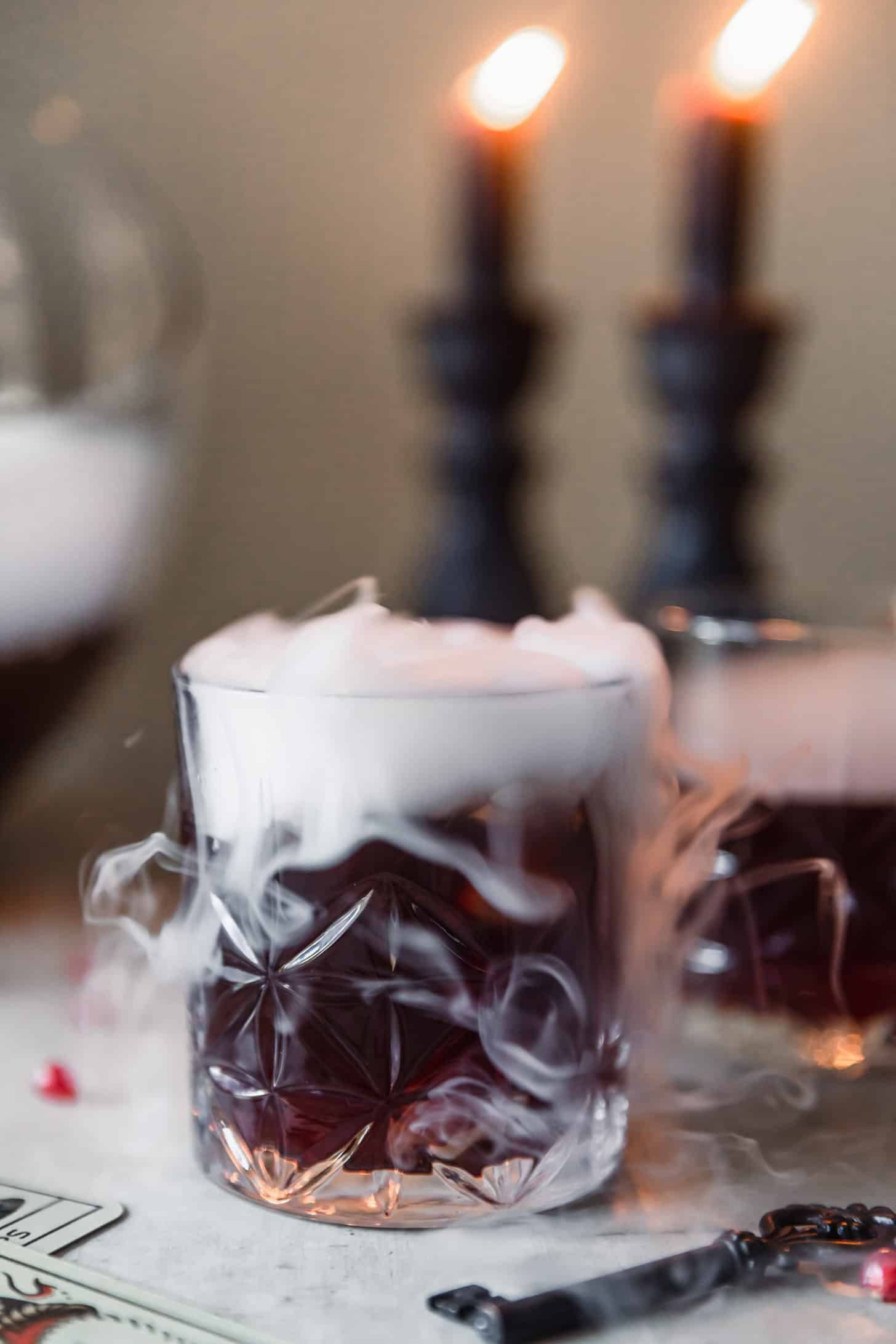 A closeup of a red-purple drink with smoke on a grey table next to vintage keys and cards.