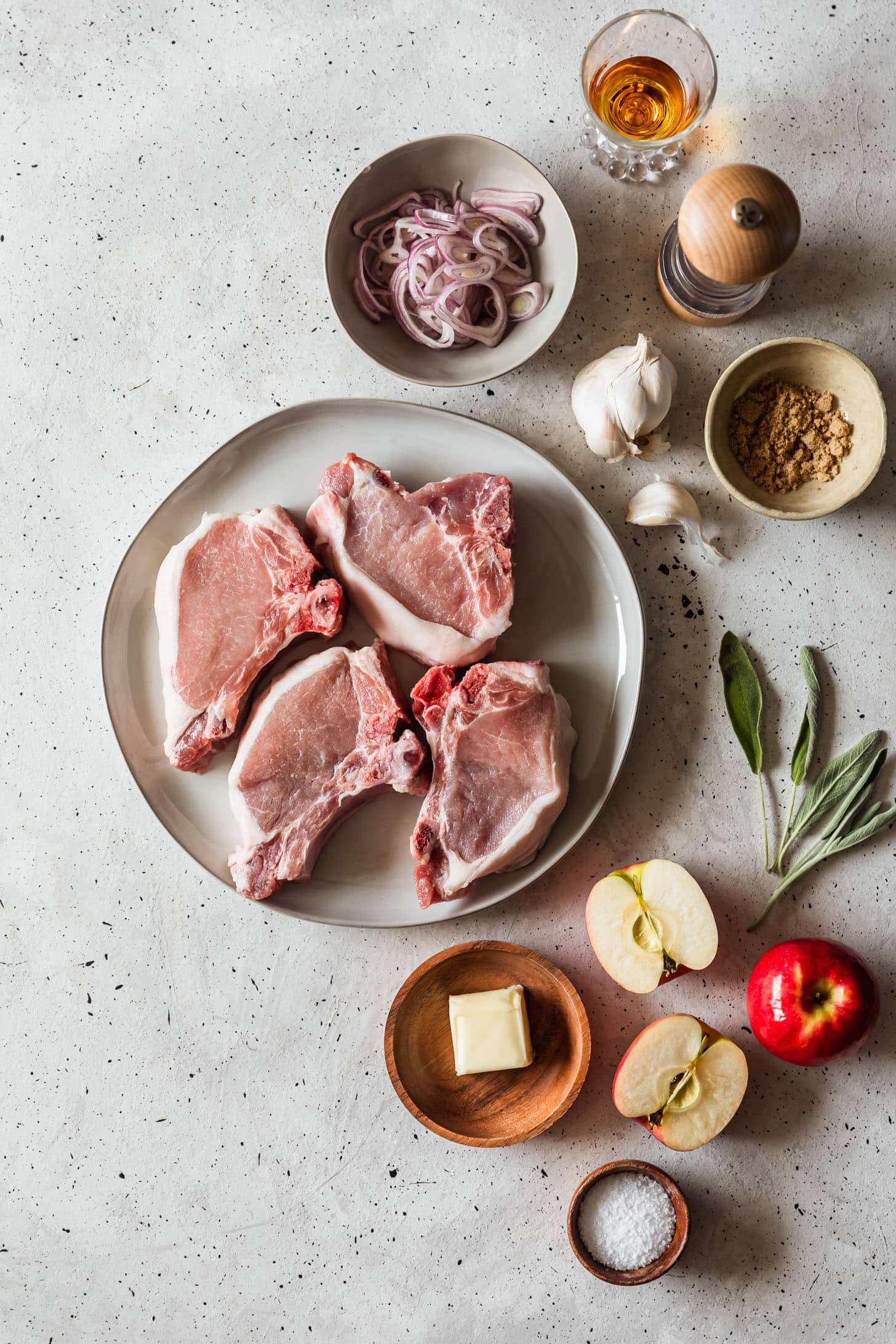 Pork chops on a white plate next to apples, sage, bourbon, garlic, brown sugar, butter, and shallots in grey and wood bowls on a white counter.