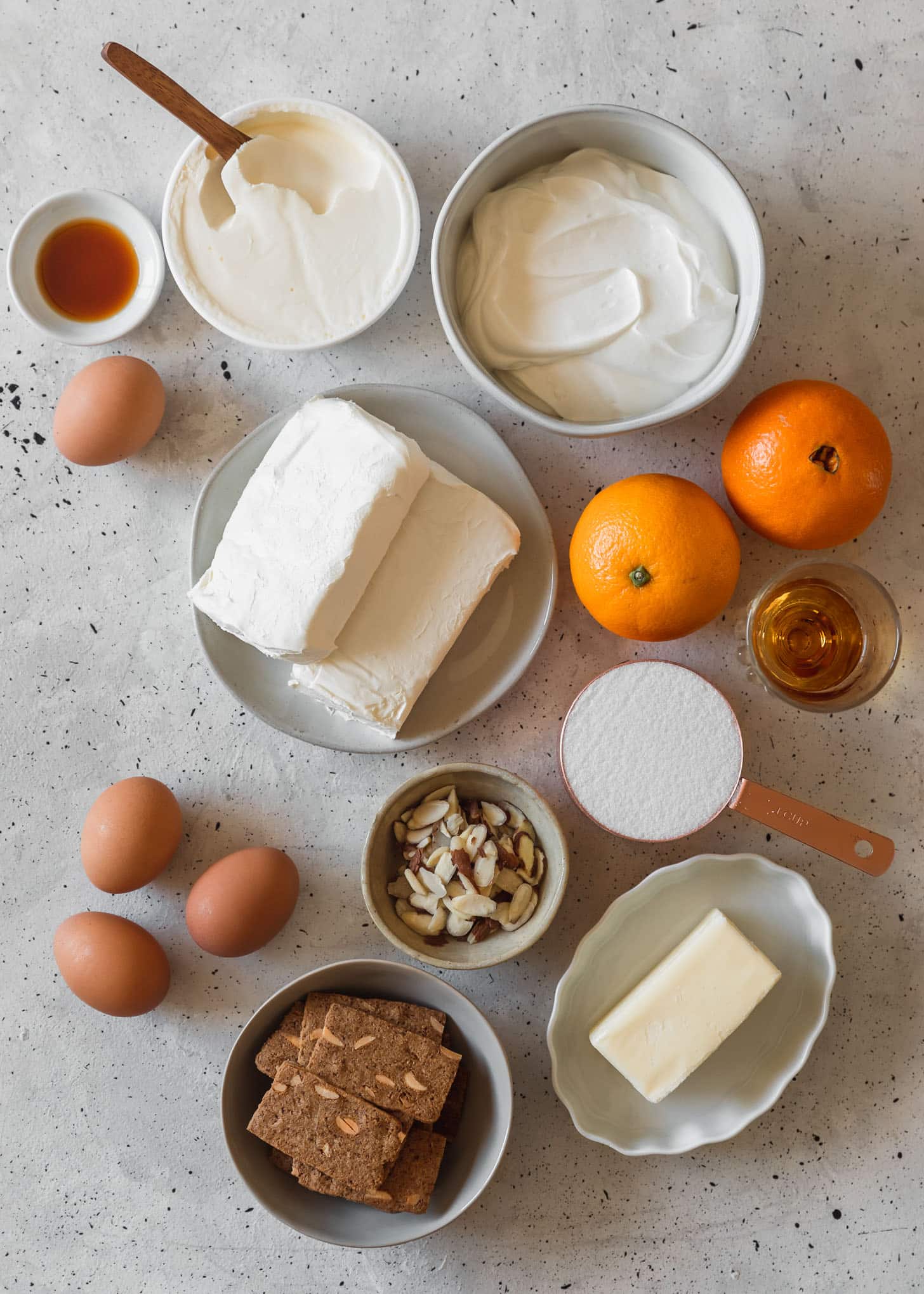 Cream cheese, mascarpone, almond cookies, sour cream, oranges, and other baking ingredients in white bowls on a grey counter.