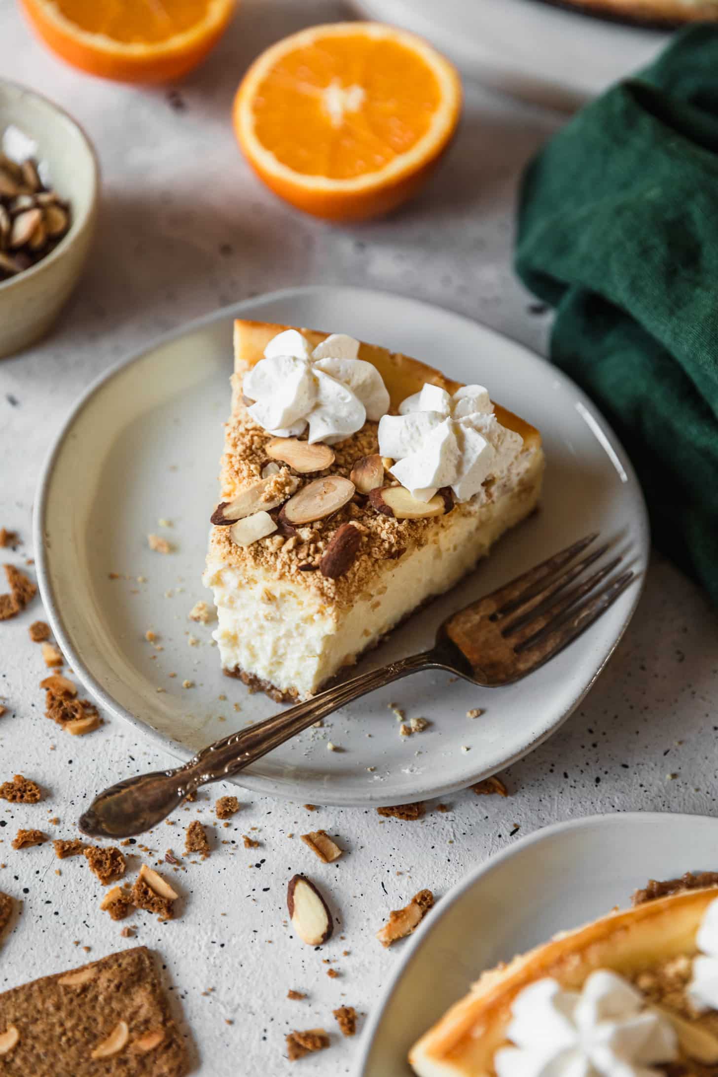 A slice of mascarpone cheesecake with a bite taken out of it on a white counter next to an emerald green napkin, sliced orange, and white bowl of almonds.