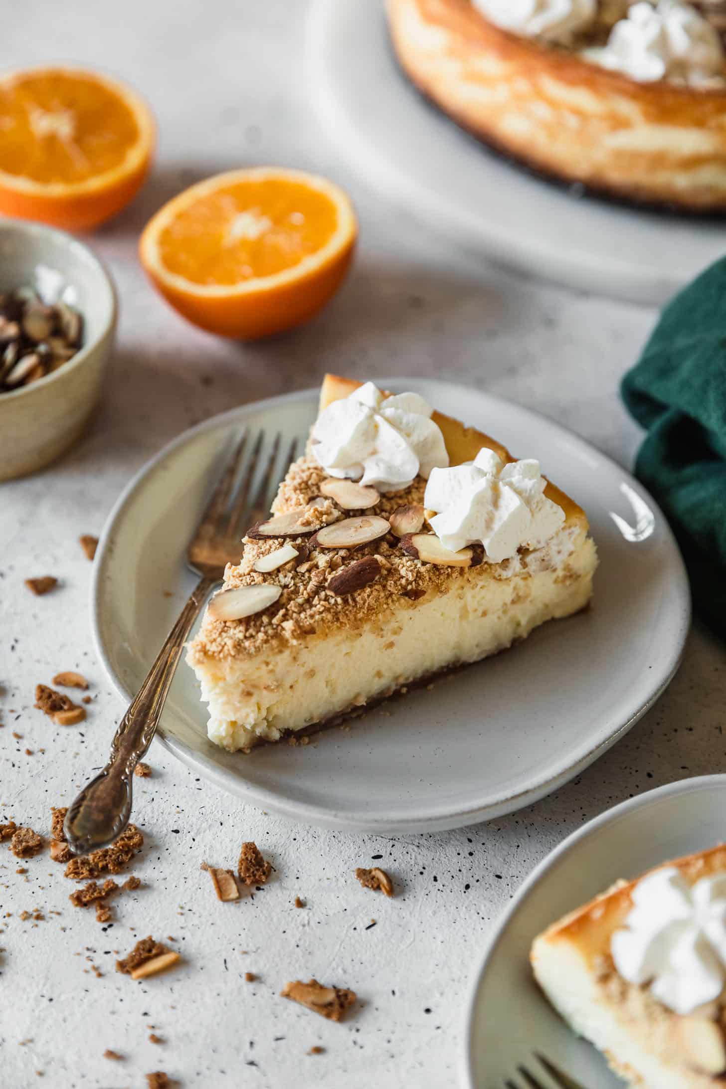 A slice of mascarpone cheesecake with orange and amaretto on a white plate next to a green napkin, oranges, and a bowl of almonds with a grey background.