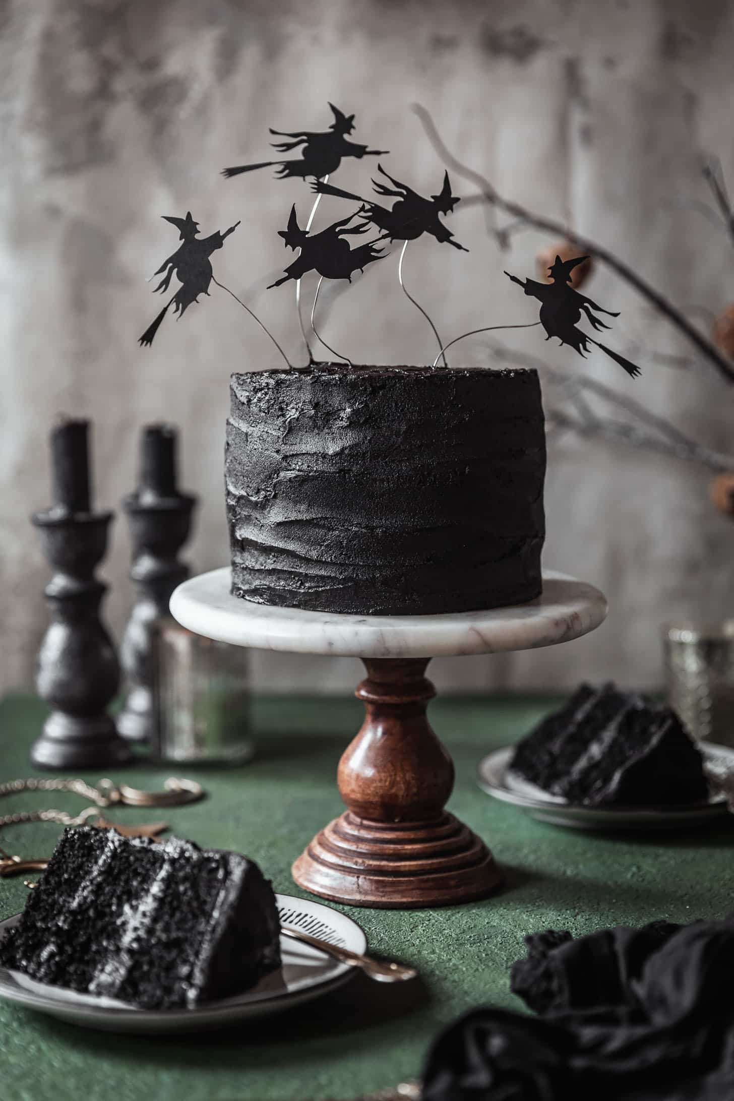 Black velvet cake with witch decorations on a marble and wood cake stand next to slices of cake and candles with a grey and dark green background.