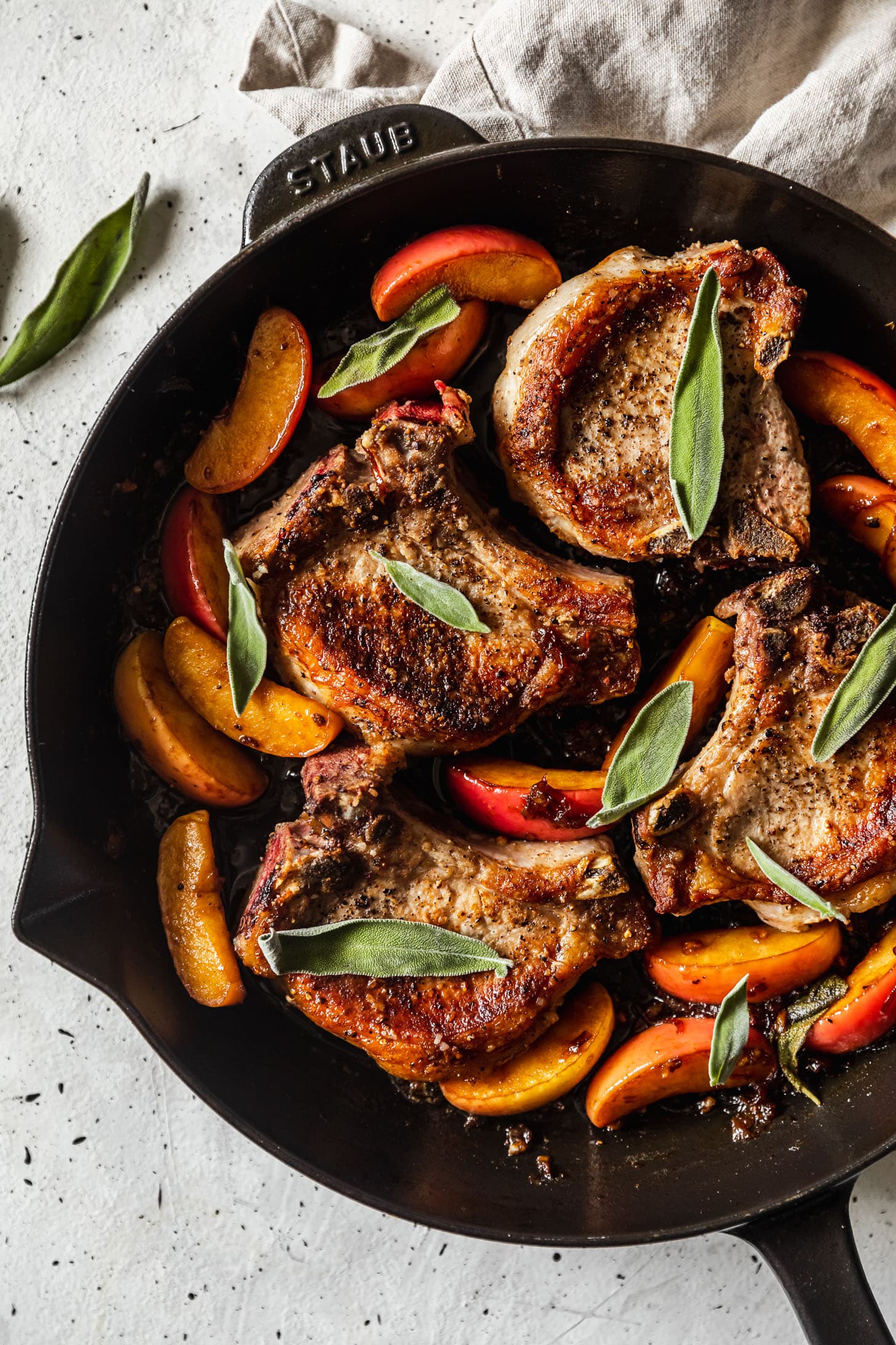 Apple pork chops with fresh sage in a black pan on a grey counter.