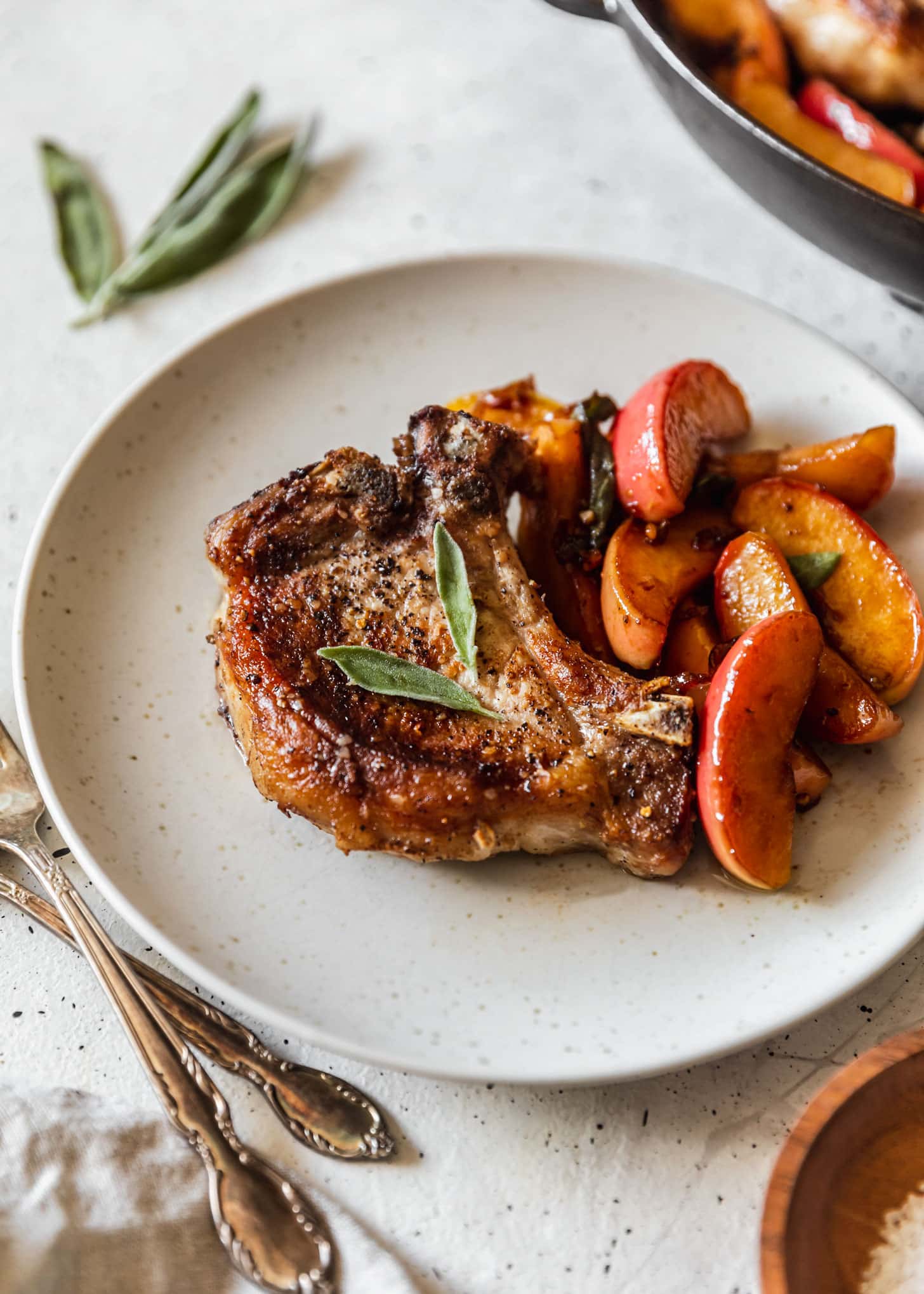 A side image of a pork chop with apples and sage on a white plate with a pan of apple pork chops in the background on a white counter.