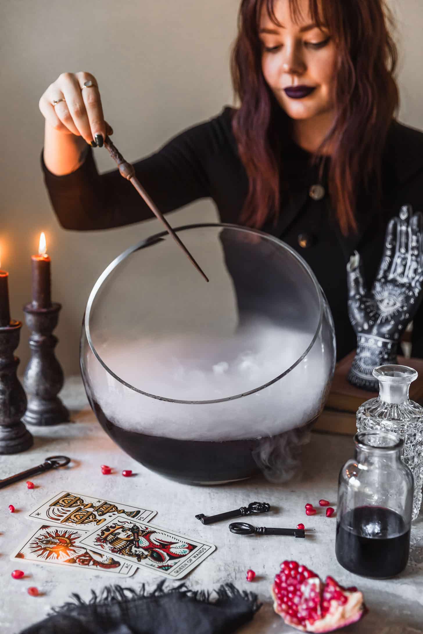 A woman in a black dress waving a wand over a bowl of witches brew cocktail punch next to potion bottles, vintage keys, and tarot cards on a grey background.