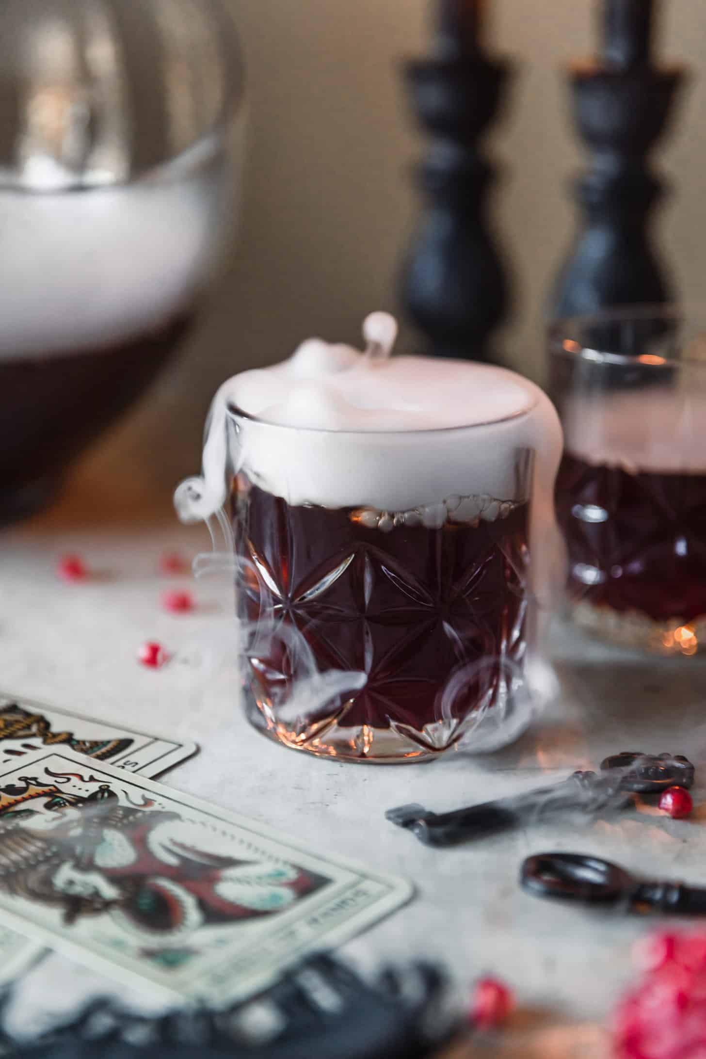 A glass of witches brew cocktail punch with dry ice on a grey counter next to a punch bowl, black candles, tarot cards, and pomegranate arils.