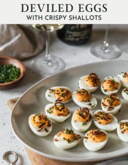 Spicy deviled eggs on a white tray next to wine glasses and chopped chives on a white counter.