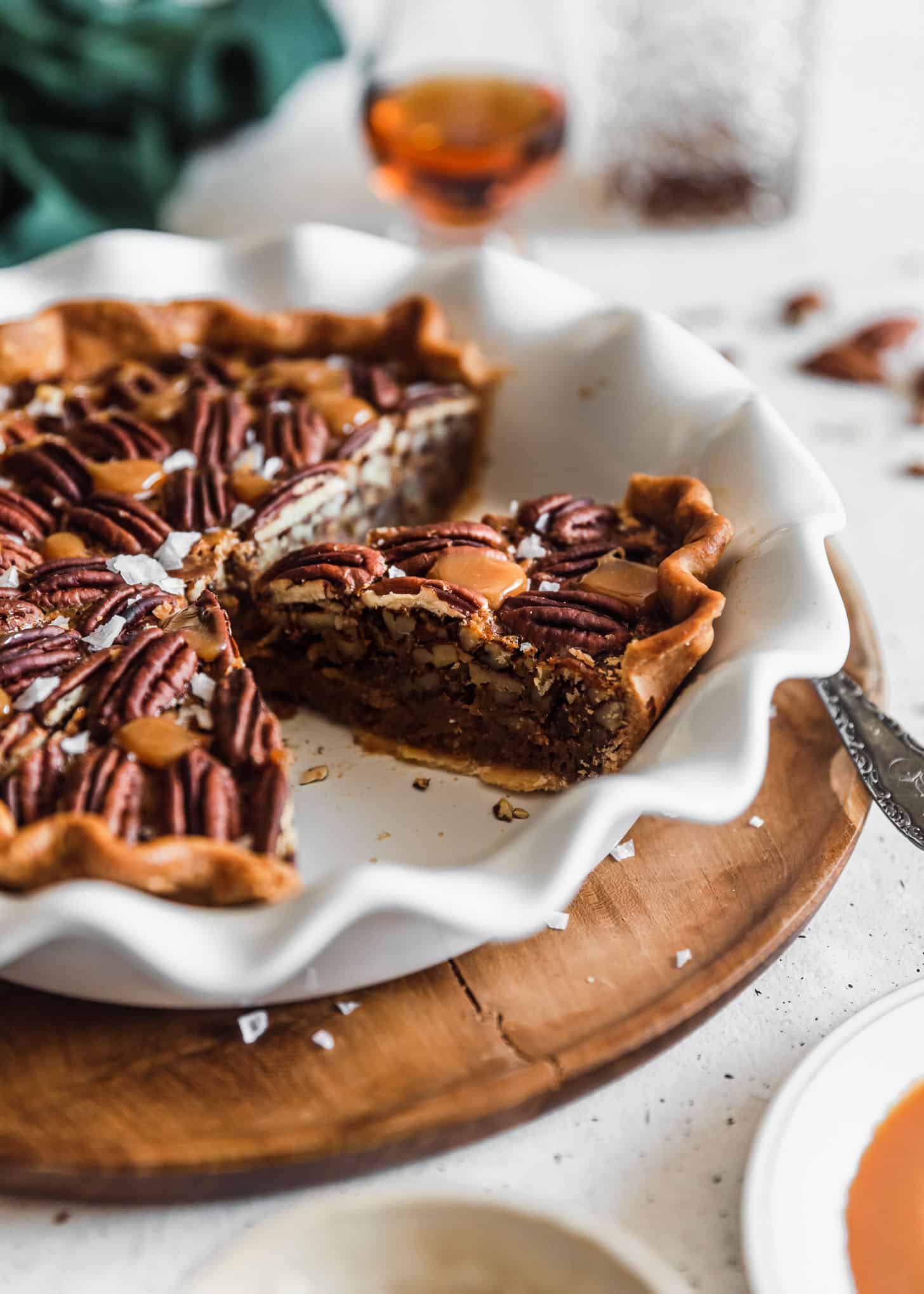 A white pie plate with slices of bourbon caramel pecan pie on a white counter with a green napkin, bourbon, and pecans in the background.