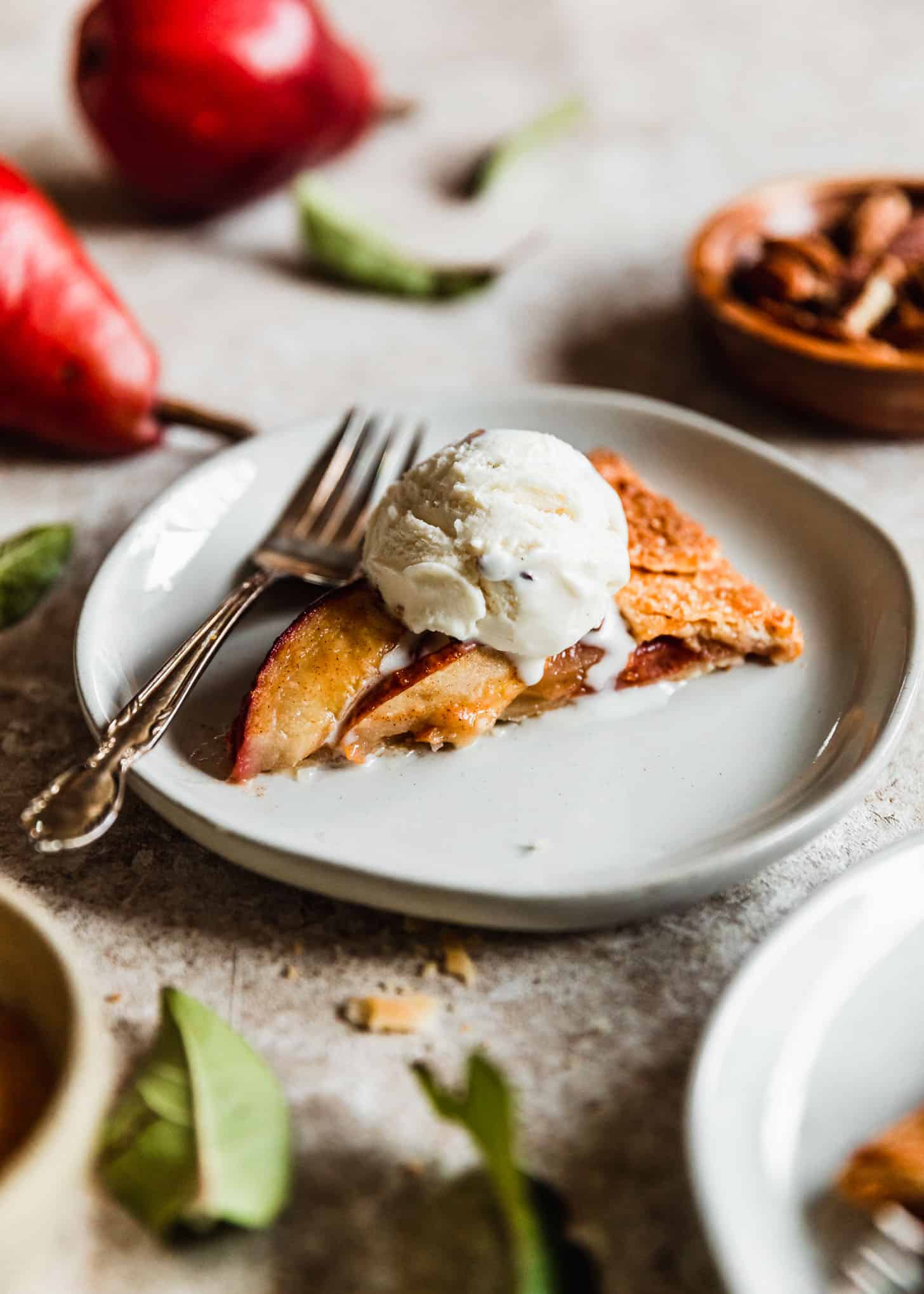 A slice of ginger pear galette with ice cream on a white plate next to red pears and leaves on a beige counter.