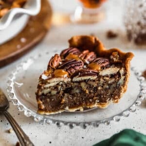 A slice of bourbon caramel pecan pie on a white counter next to a pie plate, bourbon, and a green napkin.