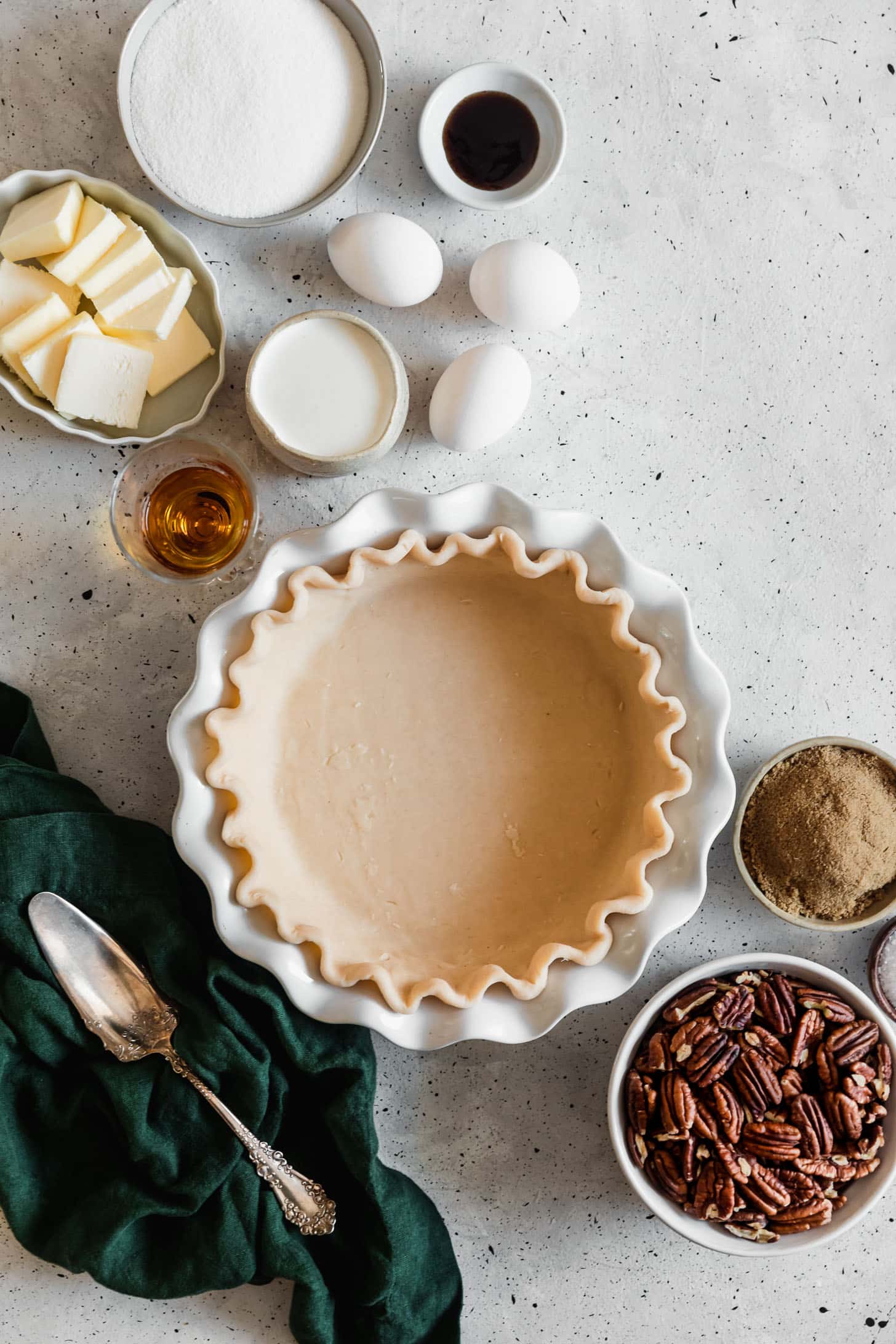 A white pie plate with dough next to white bowls of pecans, brown sugar, butter, eggs, vanilla, and bourbon on a white counter.