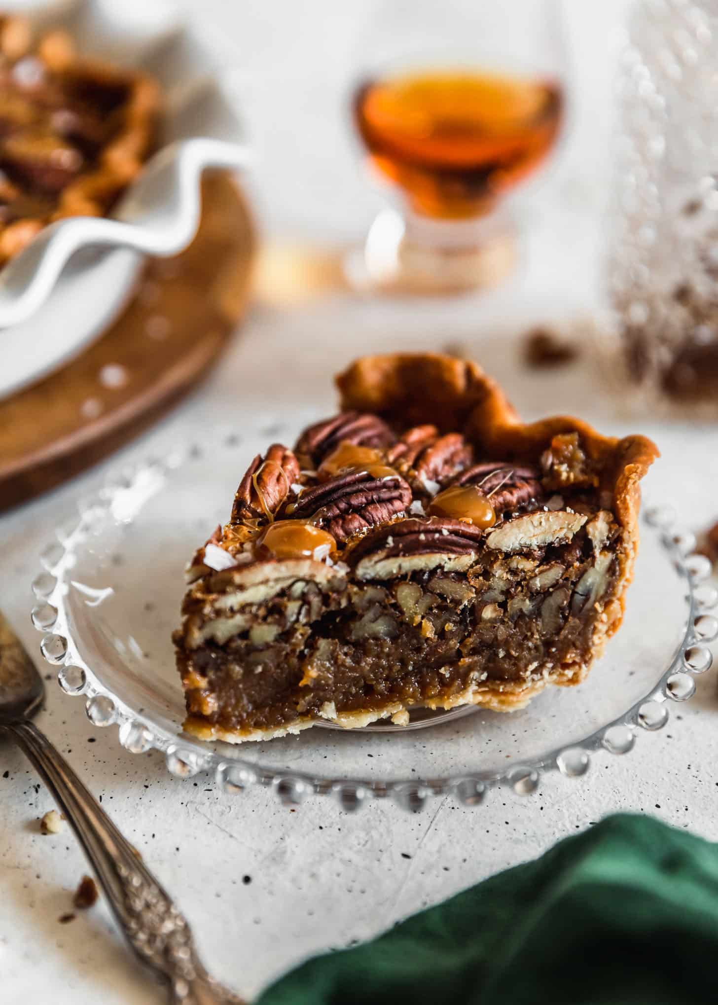 A slice of bourbon caramel pecan pie on a white table next to bourbon, a pie plate, and a green linen.