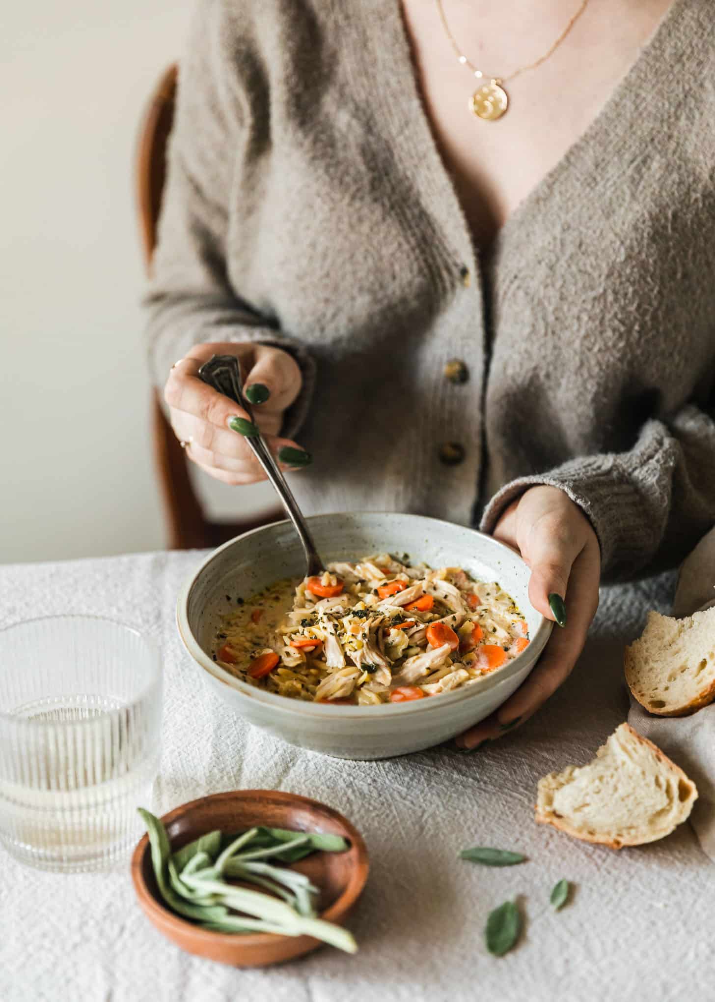 A woman in a beige sweater holding a grey bowl of creamy orzo turkey soup on a white tablecloth next to bread, white wine, and sage leaves.