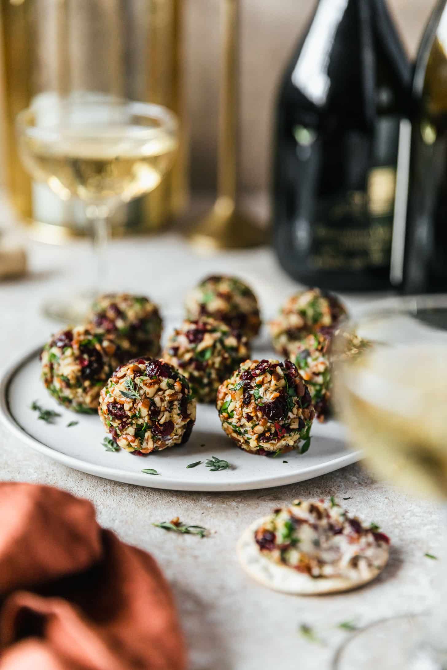 A white plate of cranberry orange cheese ball bites on a beige counter next to a cracker, glasses of champagne, and a rust colored napkin.