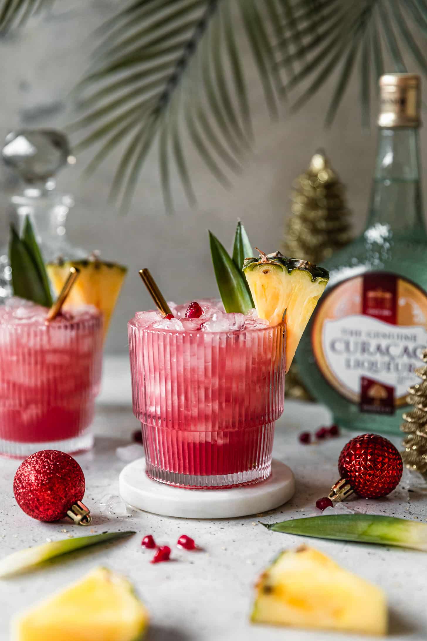 Two holiday mai tais on a white counter next to pineapple, a bottle of Curacao, and palm leaves.