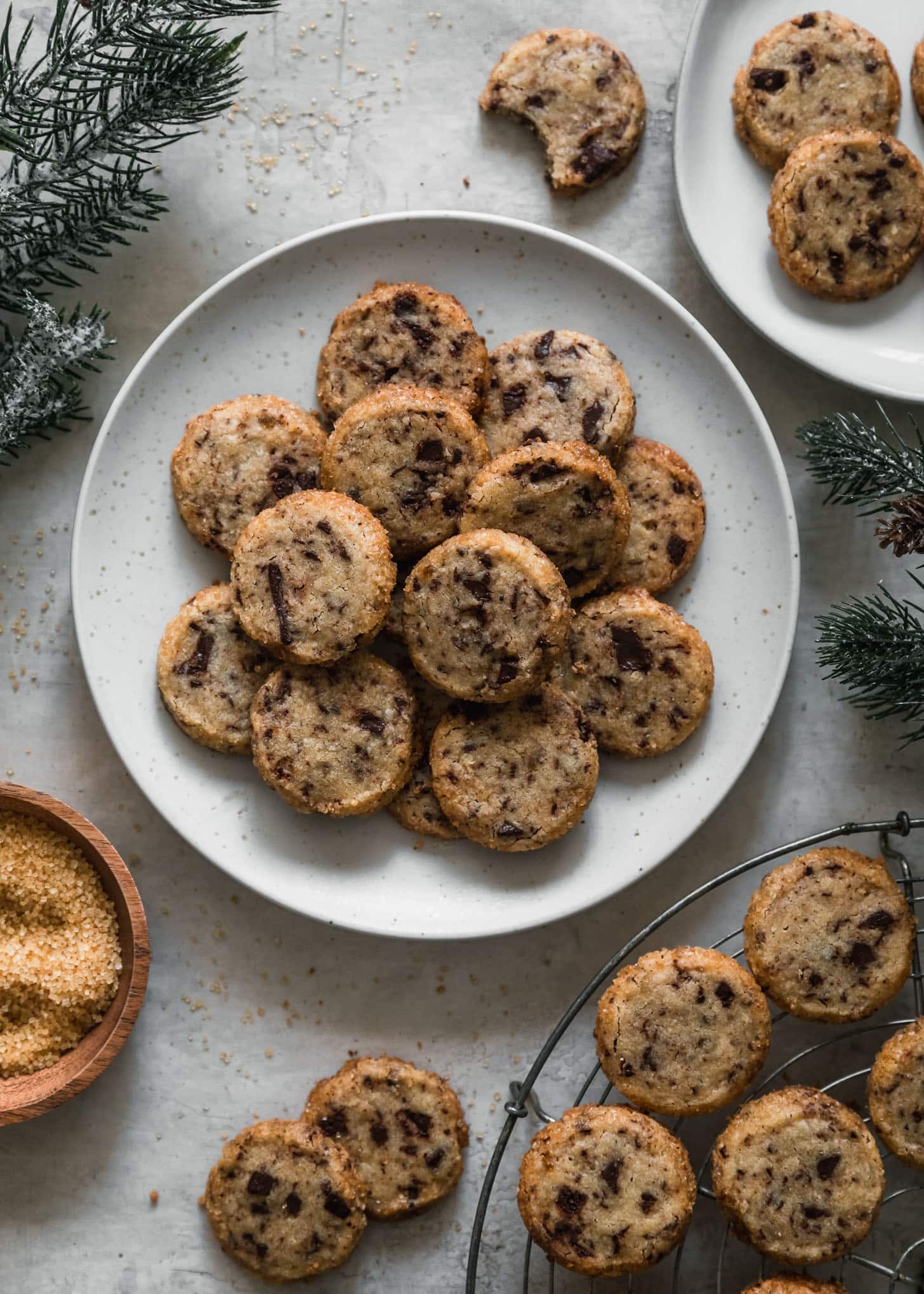 A white plate of cardamom chocolate chunk shortbread cookies next to a cooling rack, wood bowl of Turbinado sugar, and garland on a grey counter.