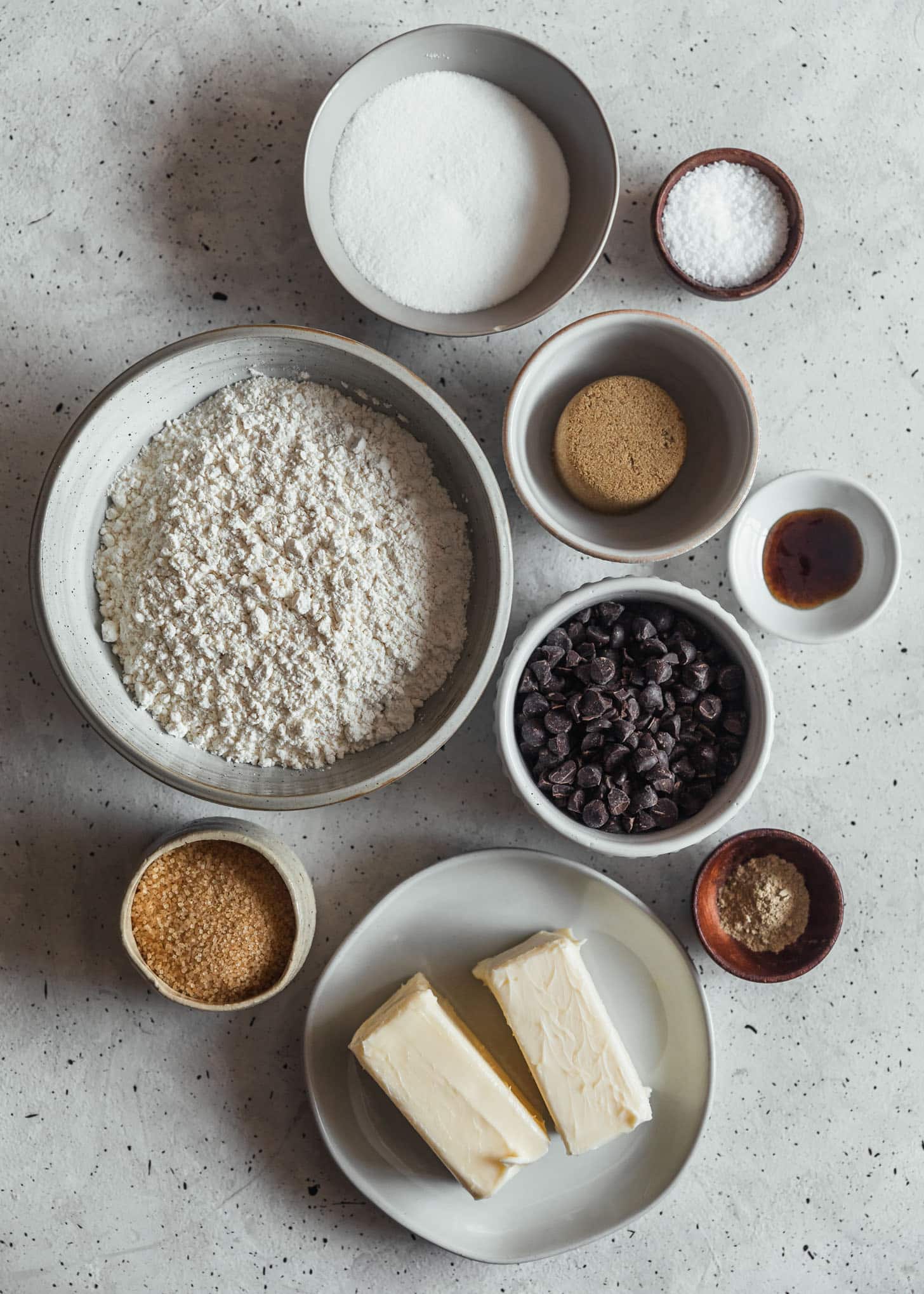 Cookie ingredients in grey, wood, and white bowls on a grey speckled counter.