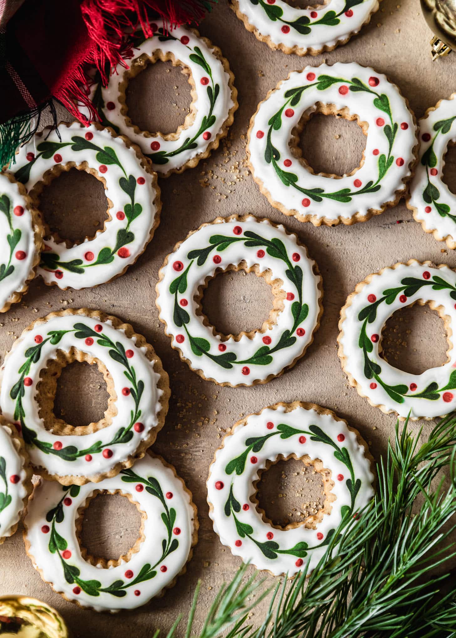 Cut out pecan sugar cookies with wreath decorations on a brown counter next to garland, a red plaid napkin, and gold ornaments.