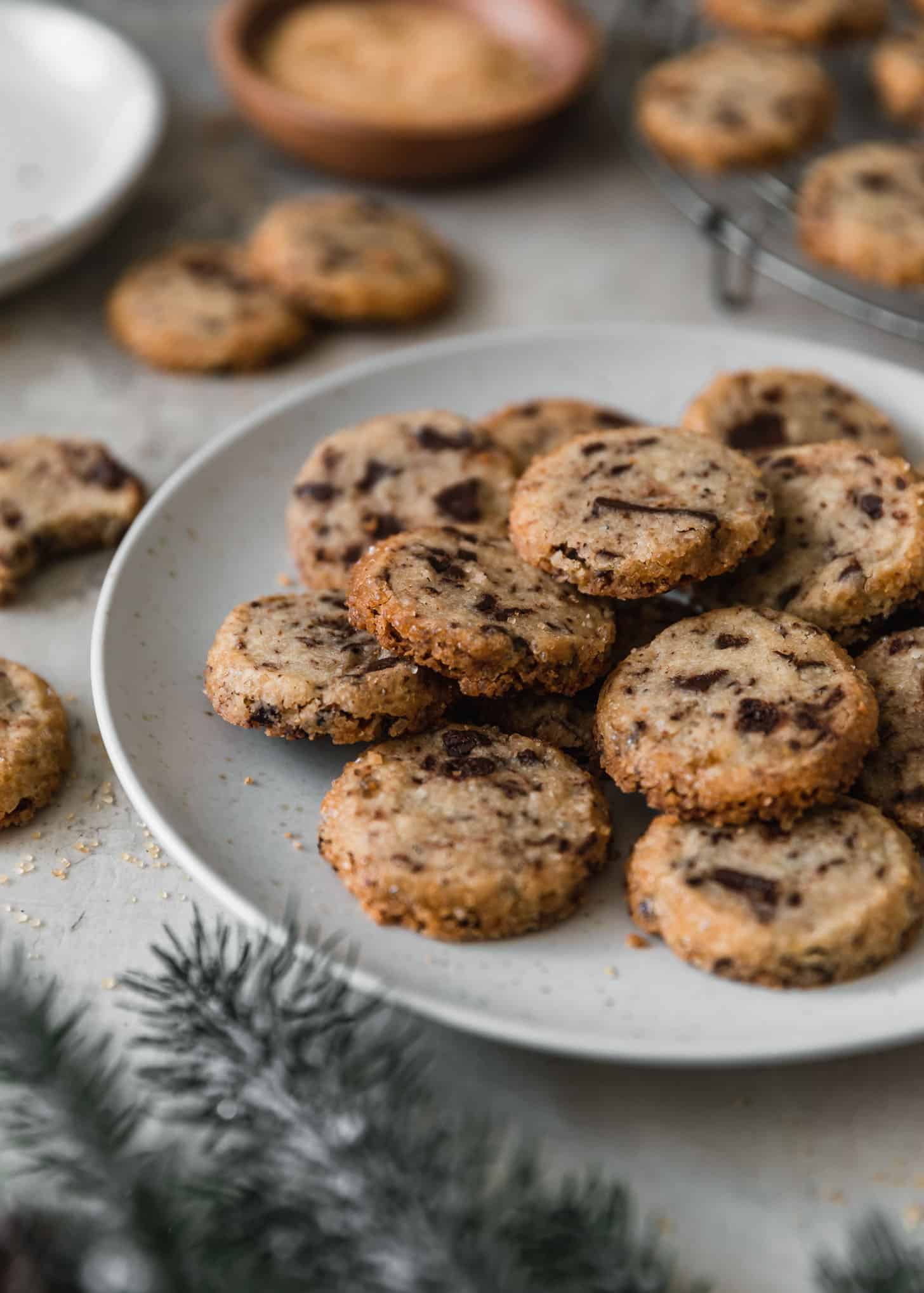 A white plate of cardamom chocolate chip shortbread cookies next to a cooling rack, garland, and more cookies with a grey background.