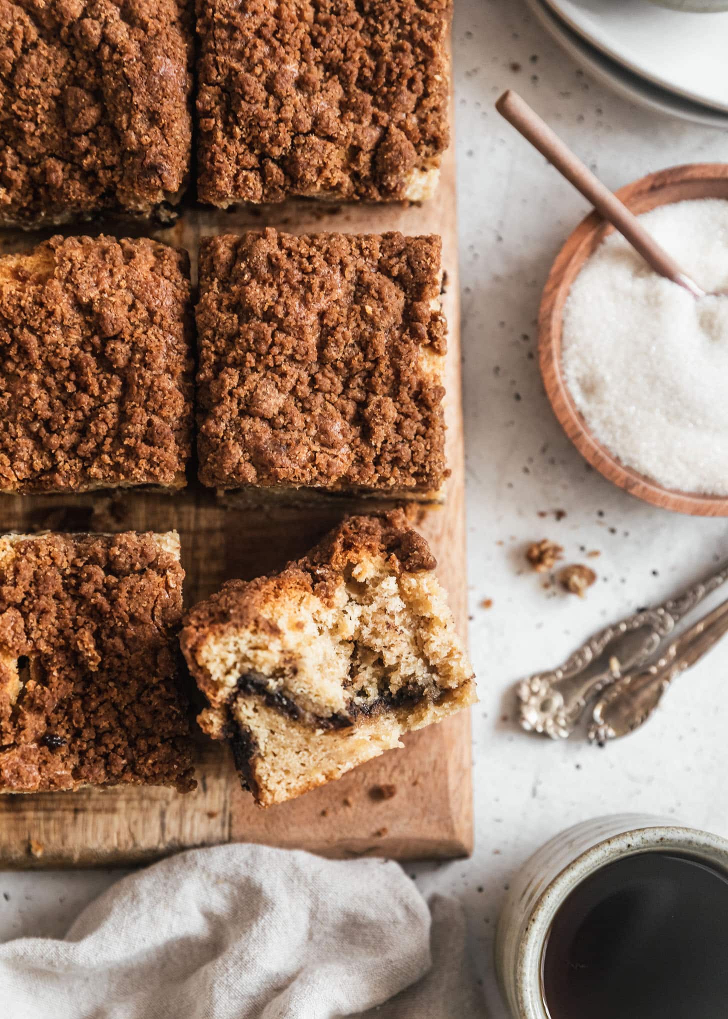 A slice of banana coffee cake with a bite taken out of it on a wood board next to more coffee cake, sugar, a cup of coffee, and forks on a white counter.