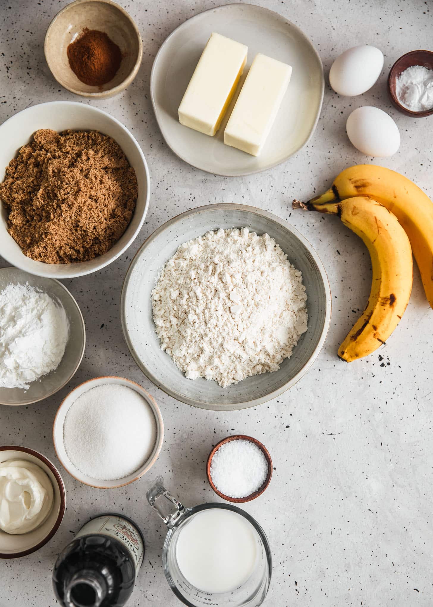 White and grey bowls of baking ingredients on a white counter next to bananas and butter.