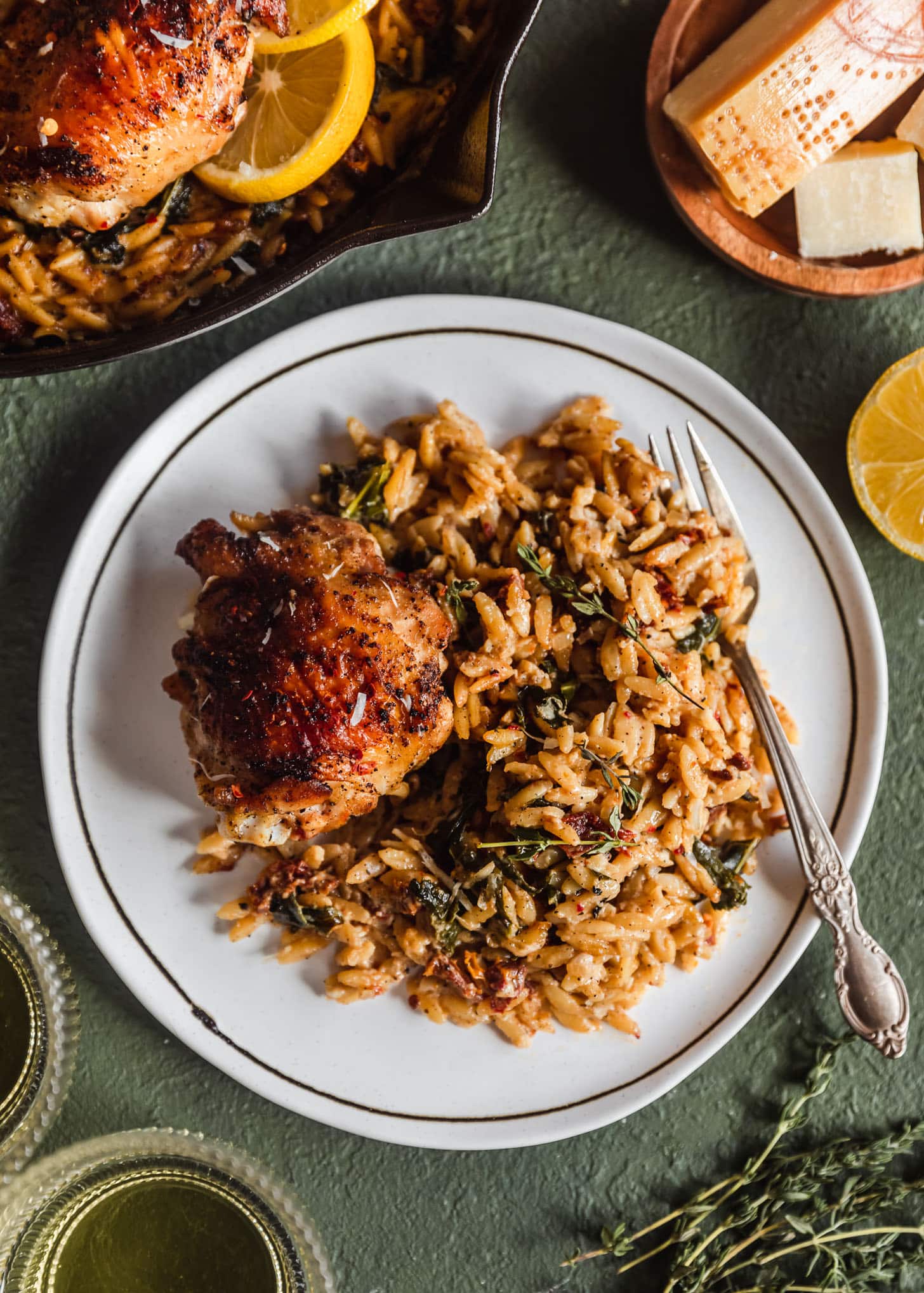 A white plate of Parmesan orzo with Tuscan chicken on a green background next to glasses of white wine, Parmesan, a skillet, and lemons.