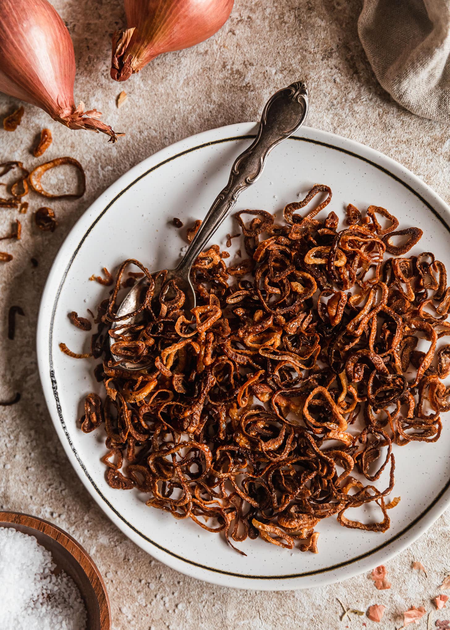 A white plate of crispy fried shallots with a spoon next to a beige linen and shallots on a tan counter.