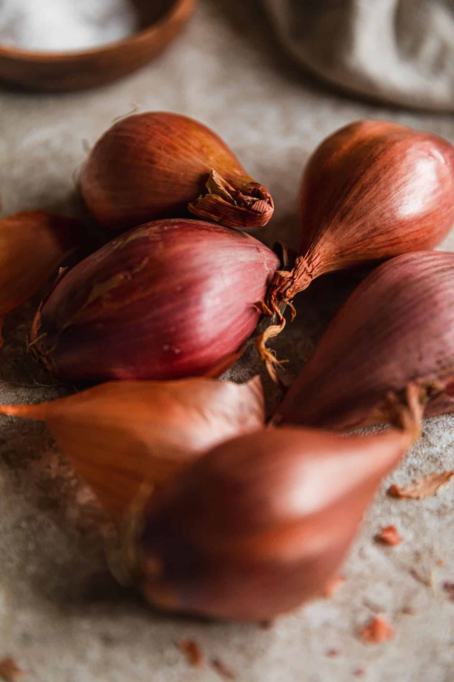 Shallots on a tan counter next to a wood bowl of salt and beige linen.