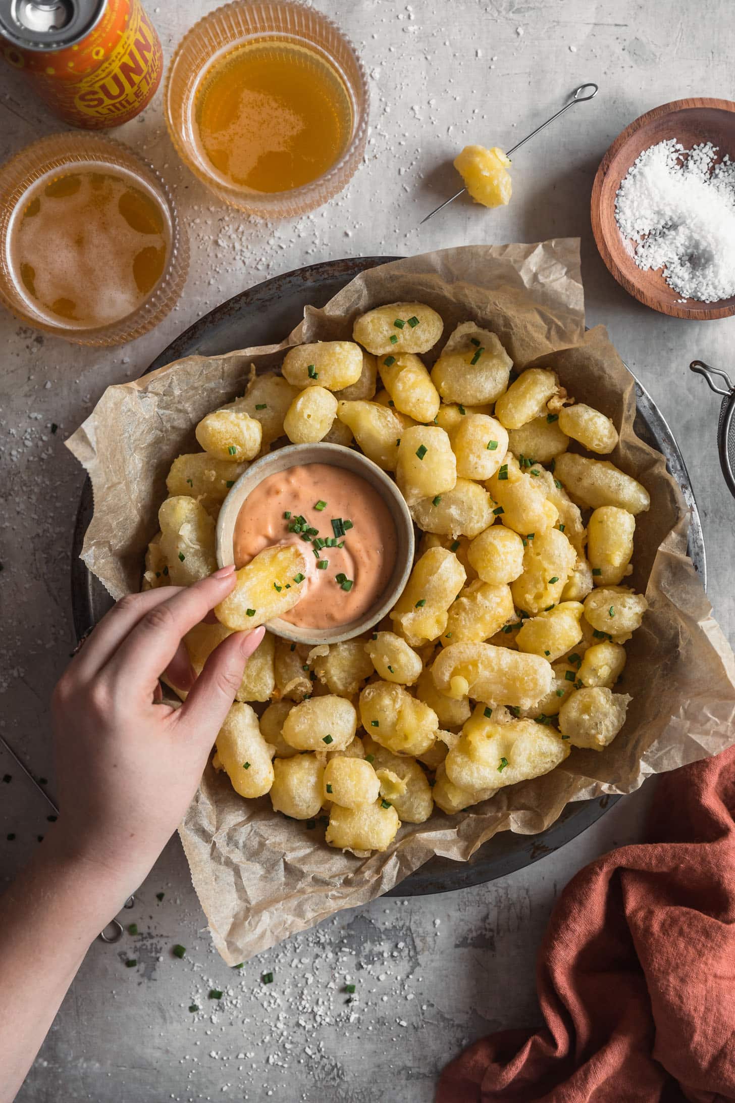 A hand dipping tempura fried cheese curds in Sriracha mayo on a plate of cheese curds next to beer and an orange linen with a grey background.