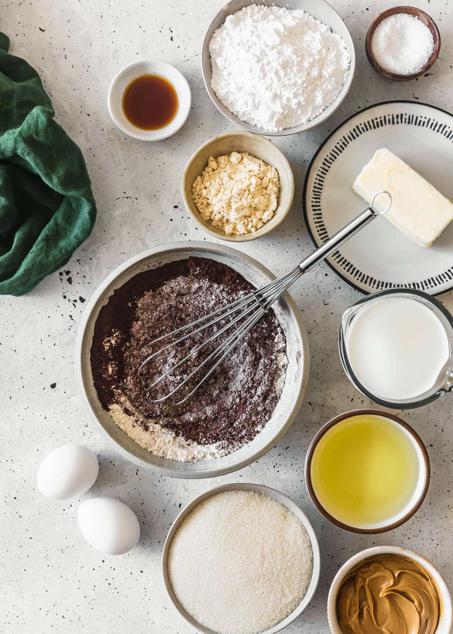 A grey bowl of flour and cocoa powder next to white bowls of sugar, nut butter, milk, milk powder, a green linen, and other baking ingredients on a white speckled background.