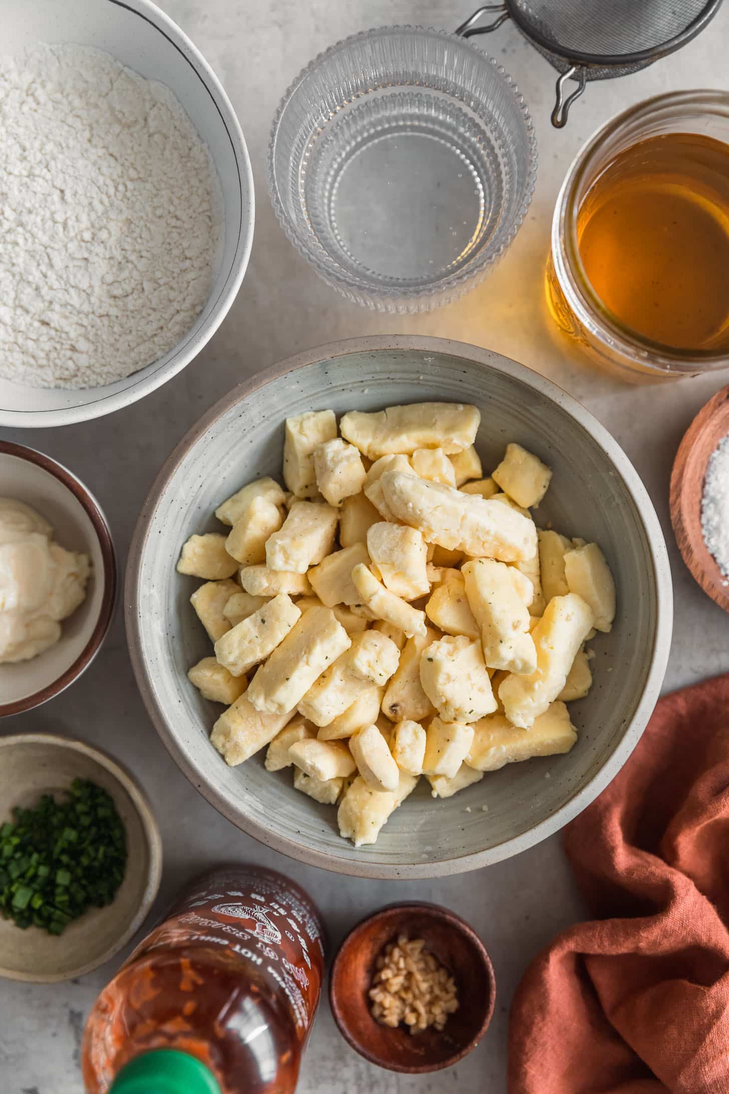 A grey bowl of cheese curds next to bowls of flour, salt, club soda, oil, and spices on a grey counter.