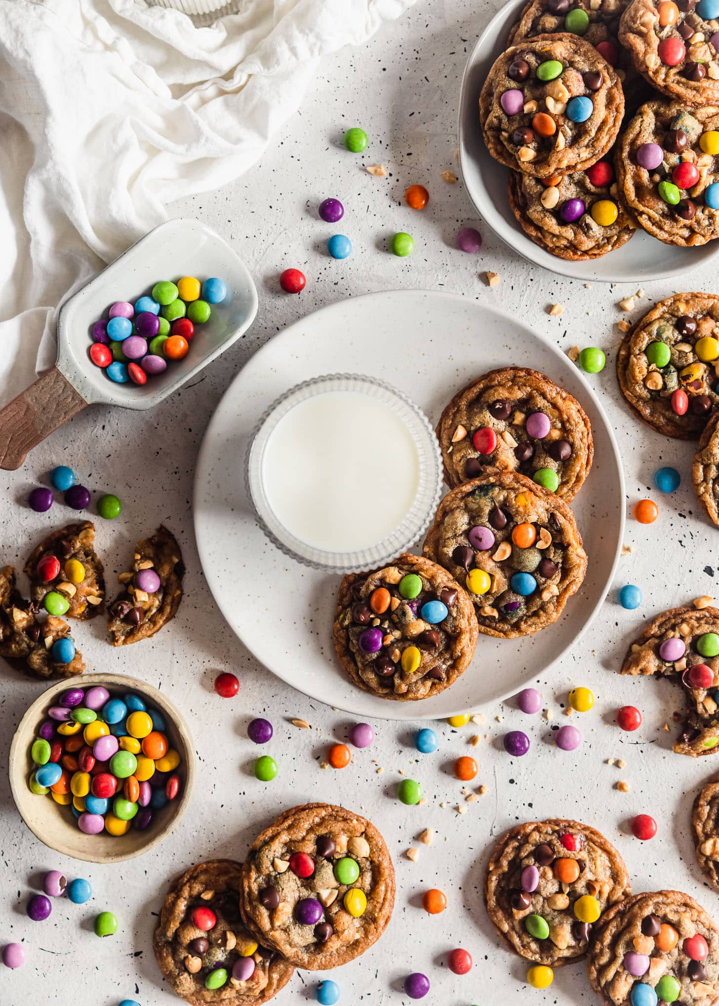 Two white plates with brown butter peanut M&M cookies next to cookies, M&Ms, glasses of milk, and a white linen on a white counter.