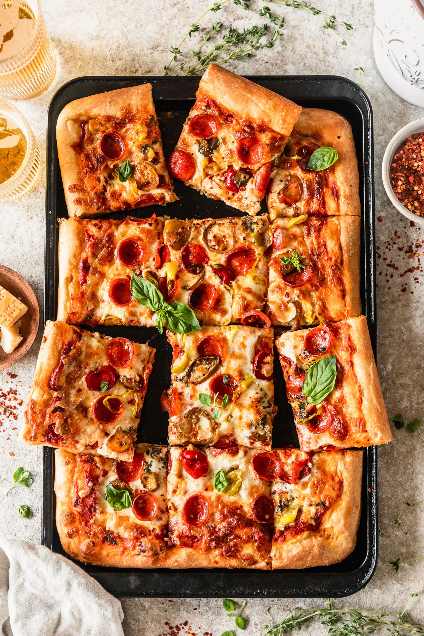 A black sheet pan with sliced Sicilian-style pizza with basil, pepperoni, and peppers on a tan counter next to a wood bowl of Parmesan, beer, and white bowl of red pepper flakes.