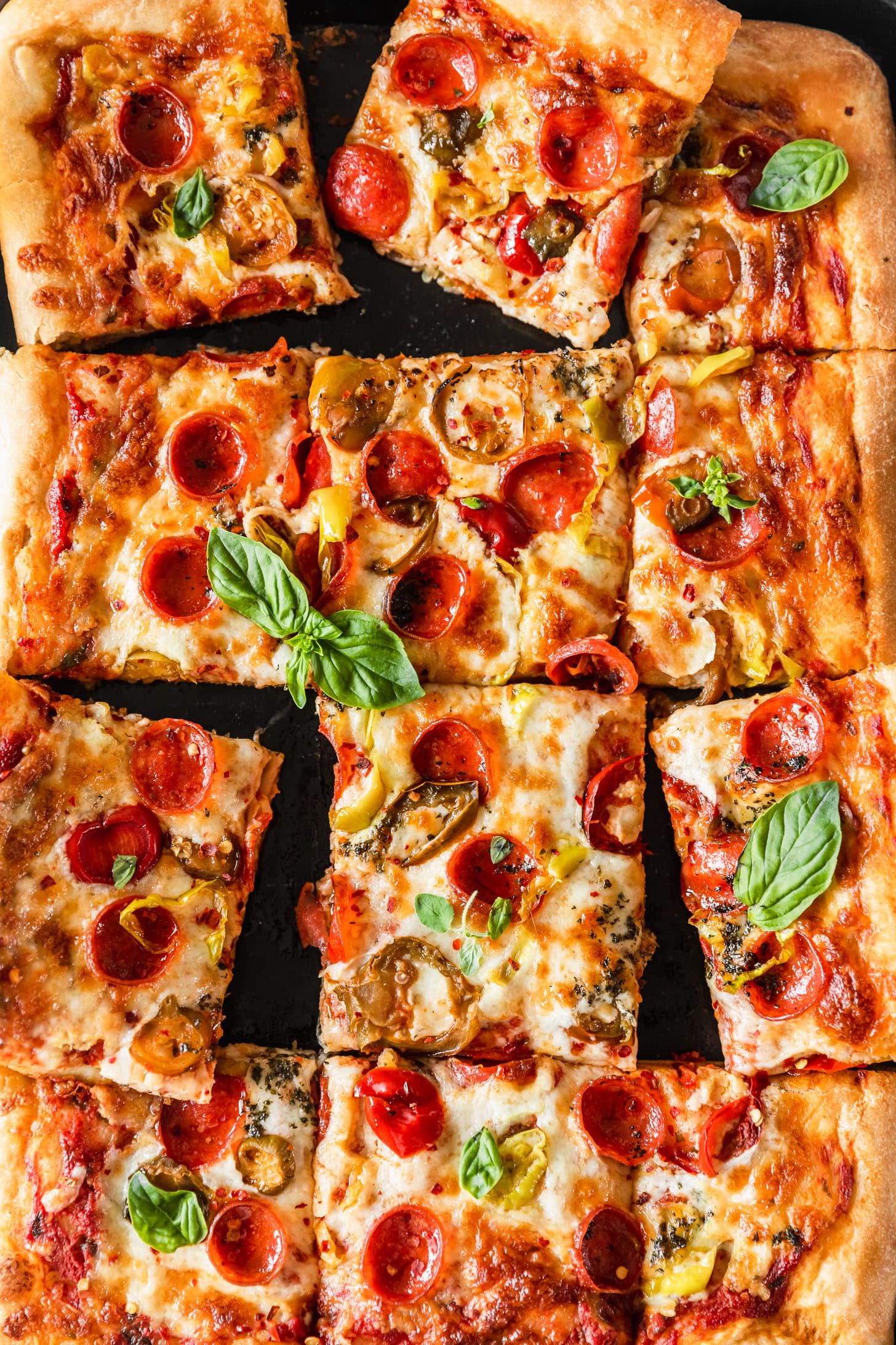 Sliced Sicilian-style pizza with pepperoni, peppers, and basil on a black sheet pan.