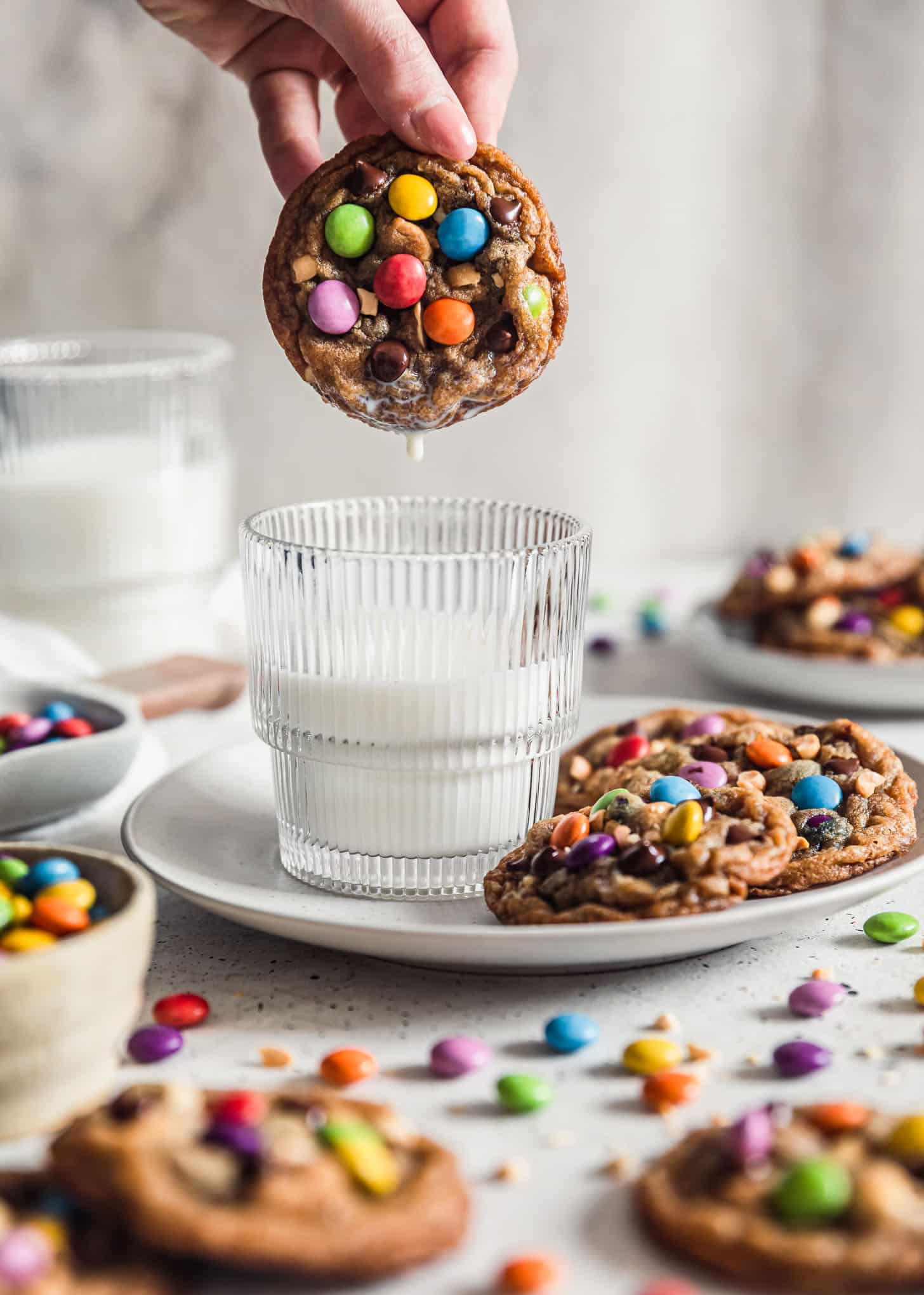 A hand dipping a brown butter peanut M&M cookie into a glass of milk on a white plate next to more cookies with a white and grey background.