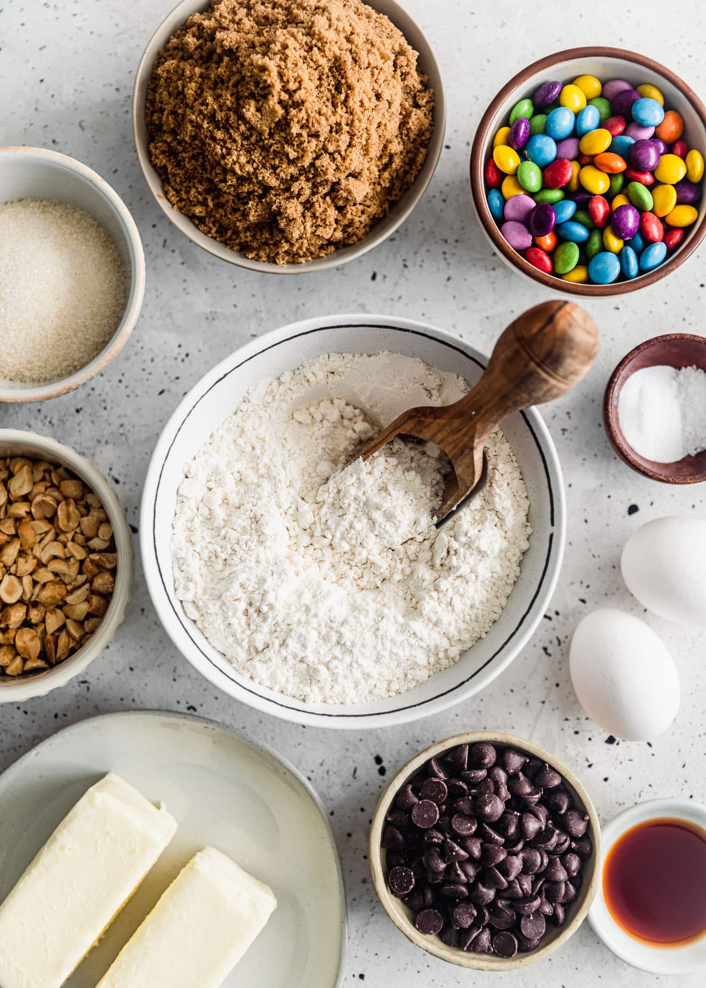 A white bowl of flour next to white bowls of flour, peanuts, sugar, brown sugar, chocolate chips, and rainbow chocolate candies on a white counter.