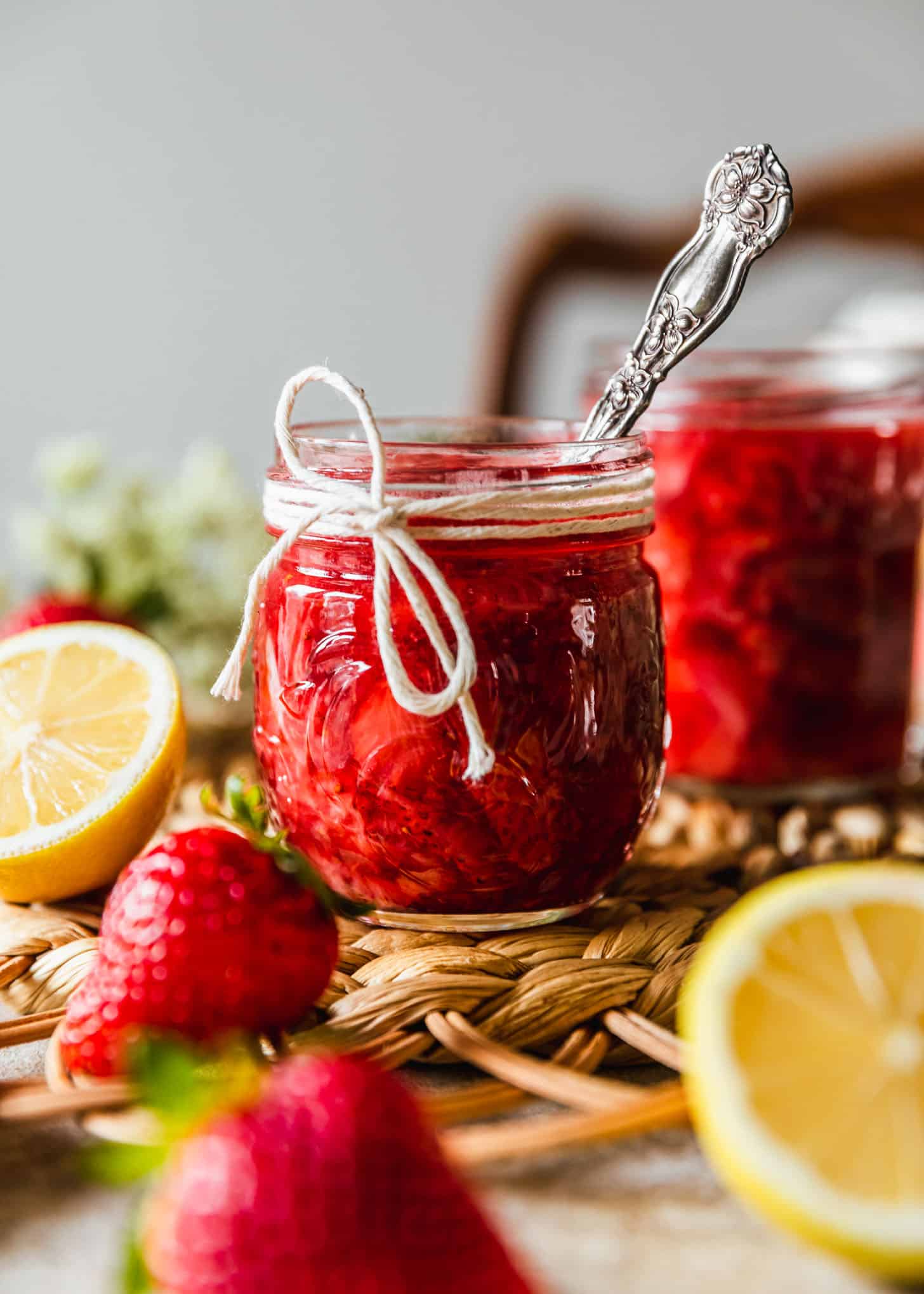A jar of strawberry compote with a spoon and a jar of strawberry coulis on a brown woven placemat next to lemons and strawberries.