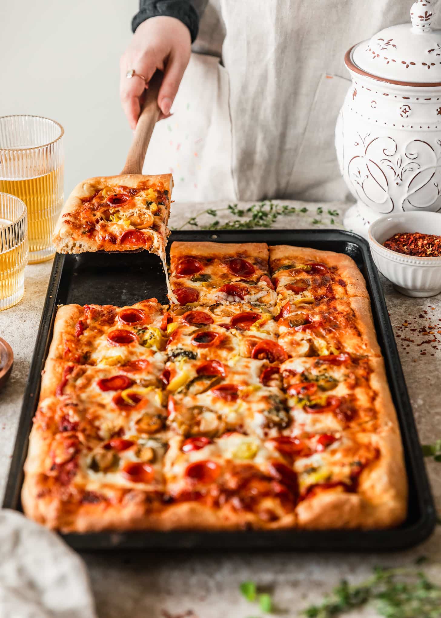 A woman wearing a tan apron using a wood spatula to pick up a piece of Sicilian-style pizza off of a sheet pan on a tan counter next to glasses of beer, thyme, a white bowl of red pepper flakes, and a white canister. 