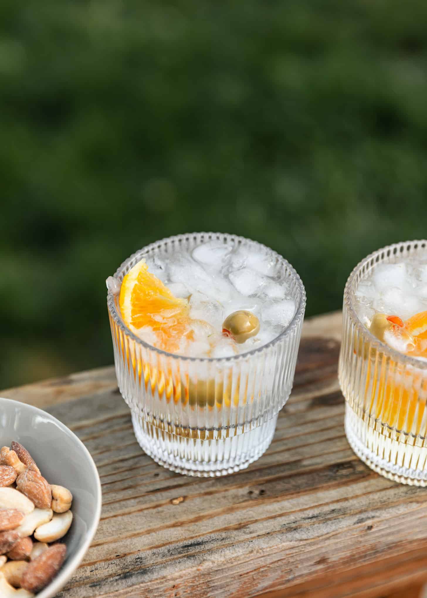 Two spritz cocktails with oranges and olives on a wood board next to a white bowl of nuts at an intimate dinner party.