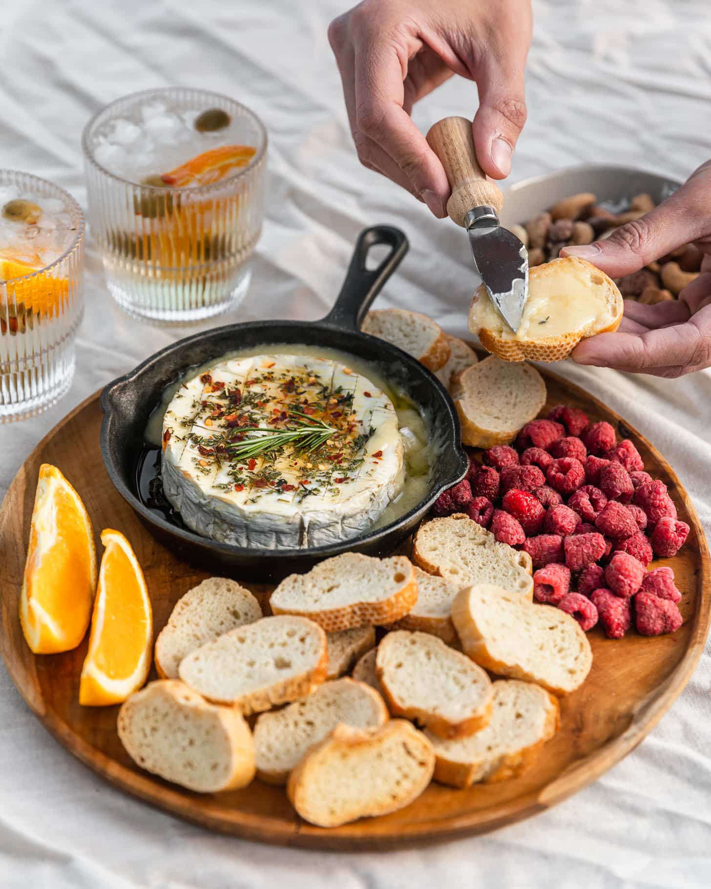 A wood board with baked Camembert, raspberries, oranges, and baguette on a white tablecloth next to a white bowl of nuts and two spritz cocktails.