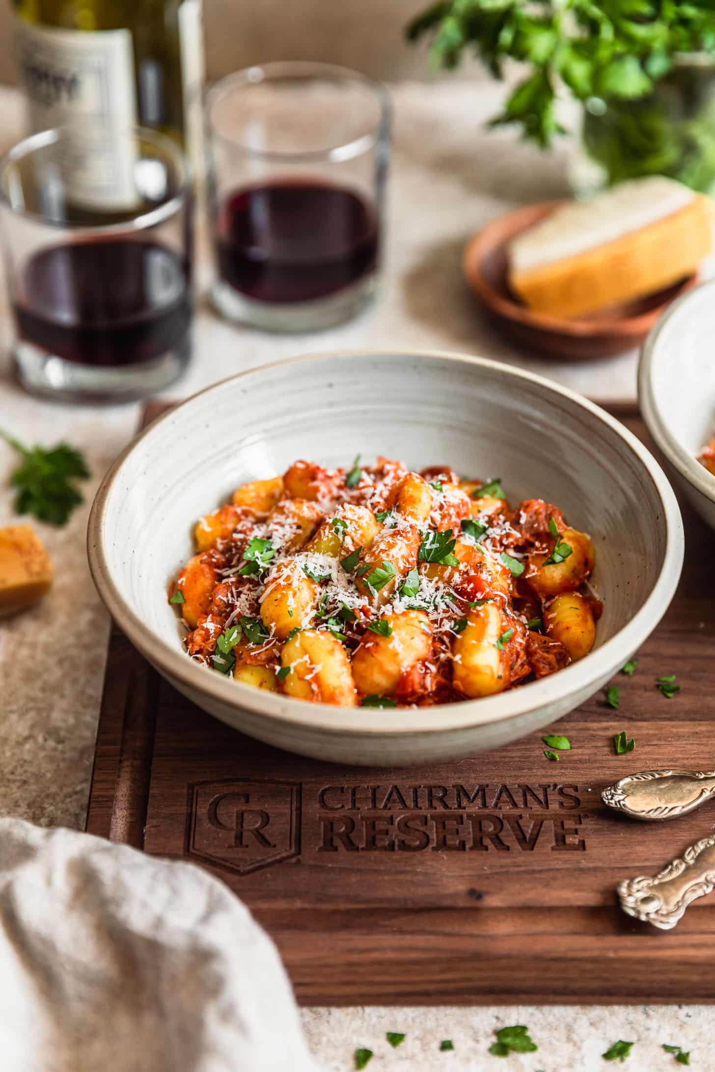 A white bowl with pork ragù with gnocchi on a wood board next to glasses of red wine, a jar of parsley, forks, and a beige linen on a tan counter.