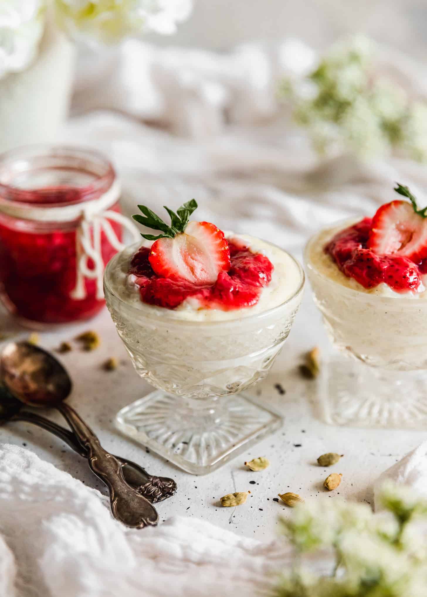 Two glass dishes of cardamom rice pudding with strawberries next to jam, spoons, cardamom pods, and white flowers on a white table.
