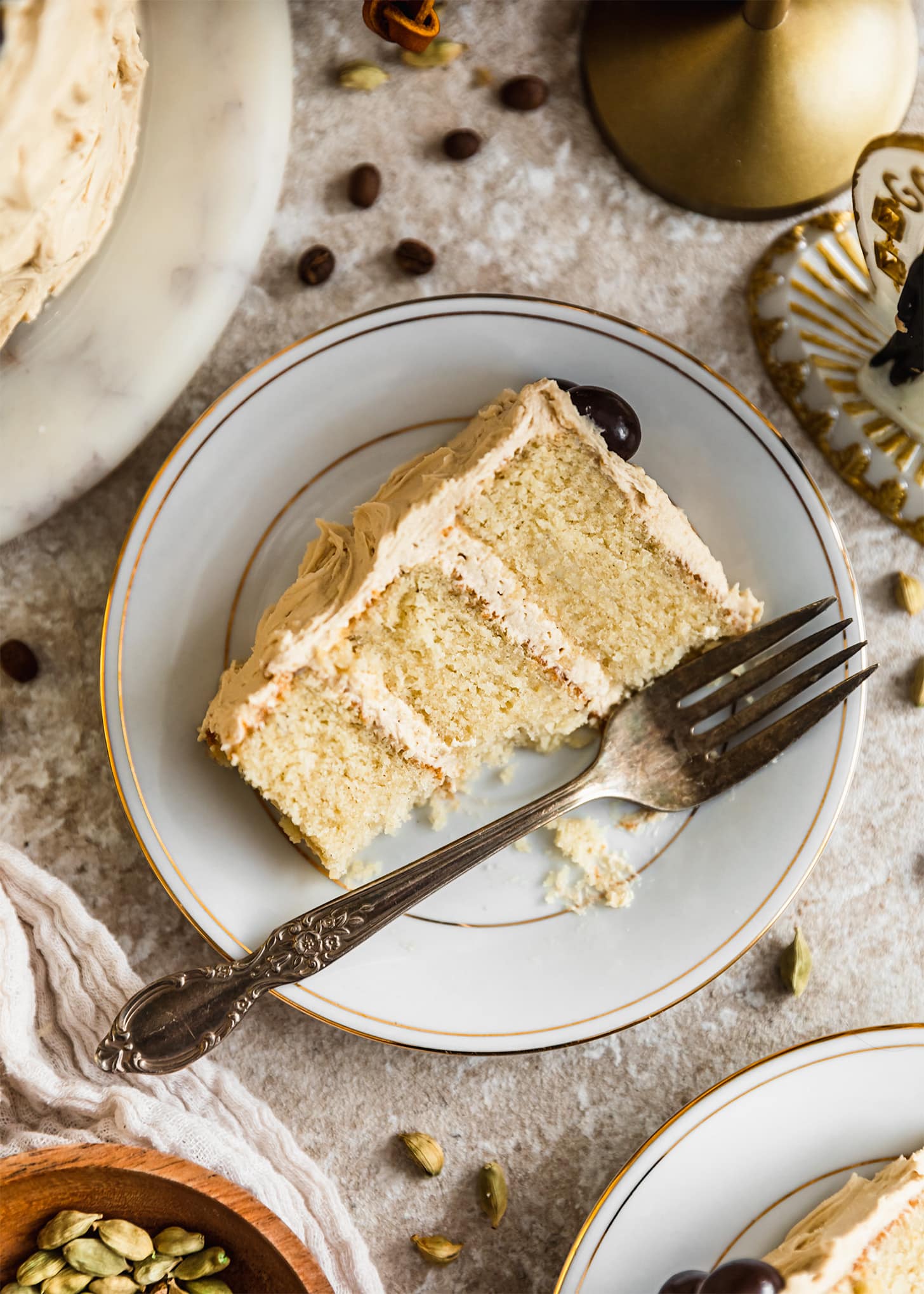 A white plate of cardamom cake with coffee frosting on a beige counter next to a white linen, wedding cake topper, coffee beans, and cardamom pods.