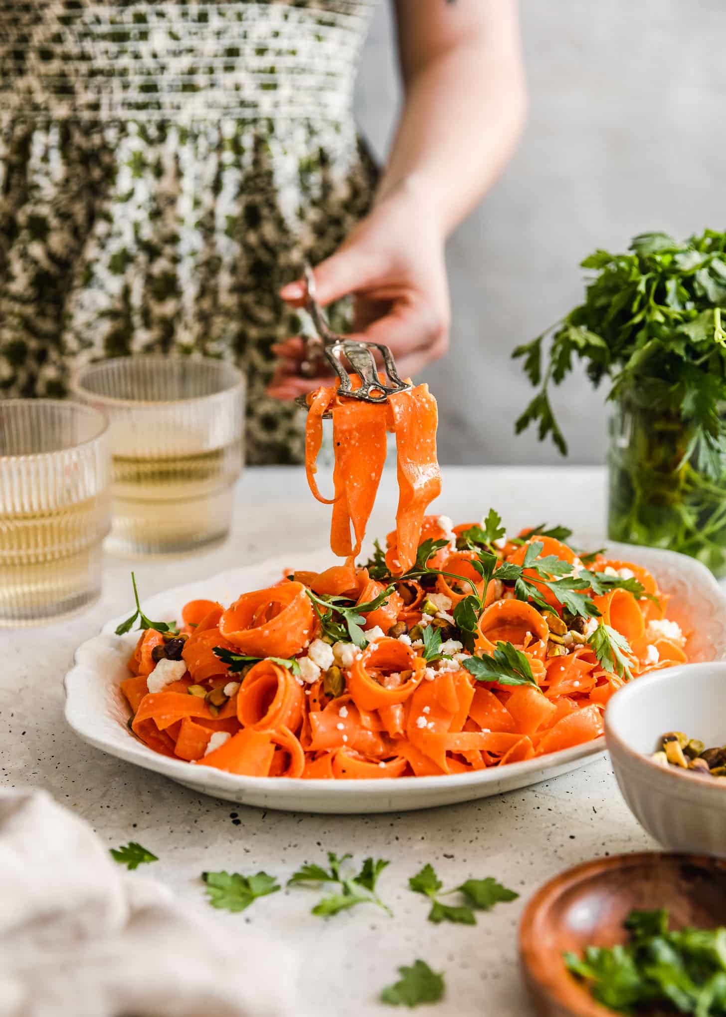 A woman in a green floral dress using tongs to grab raw carrot salad with feta, pistachios, and parsley off of a white platter next to parsley and glasses of white wine with a white and grey background.