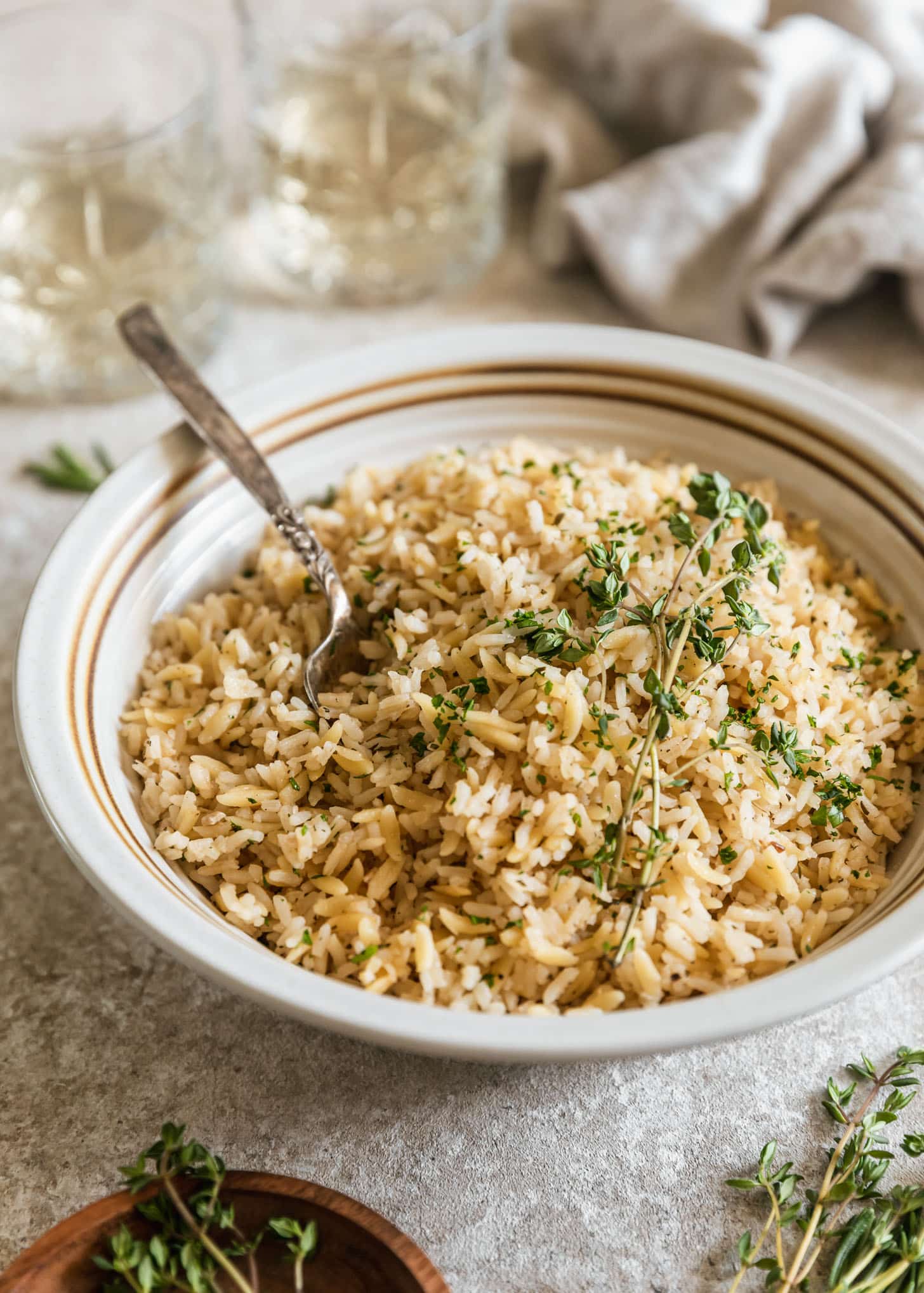 A white bowl of easy rice pilaf with orzo on a beige counter next to herbs, a beige linen, and two glasses of white wine.