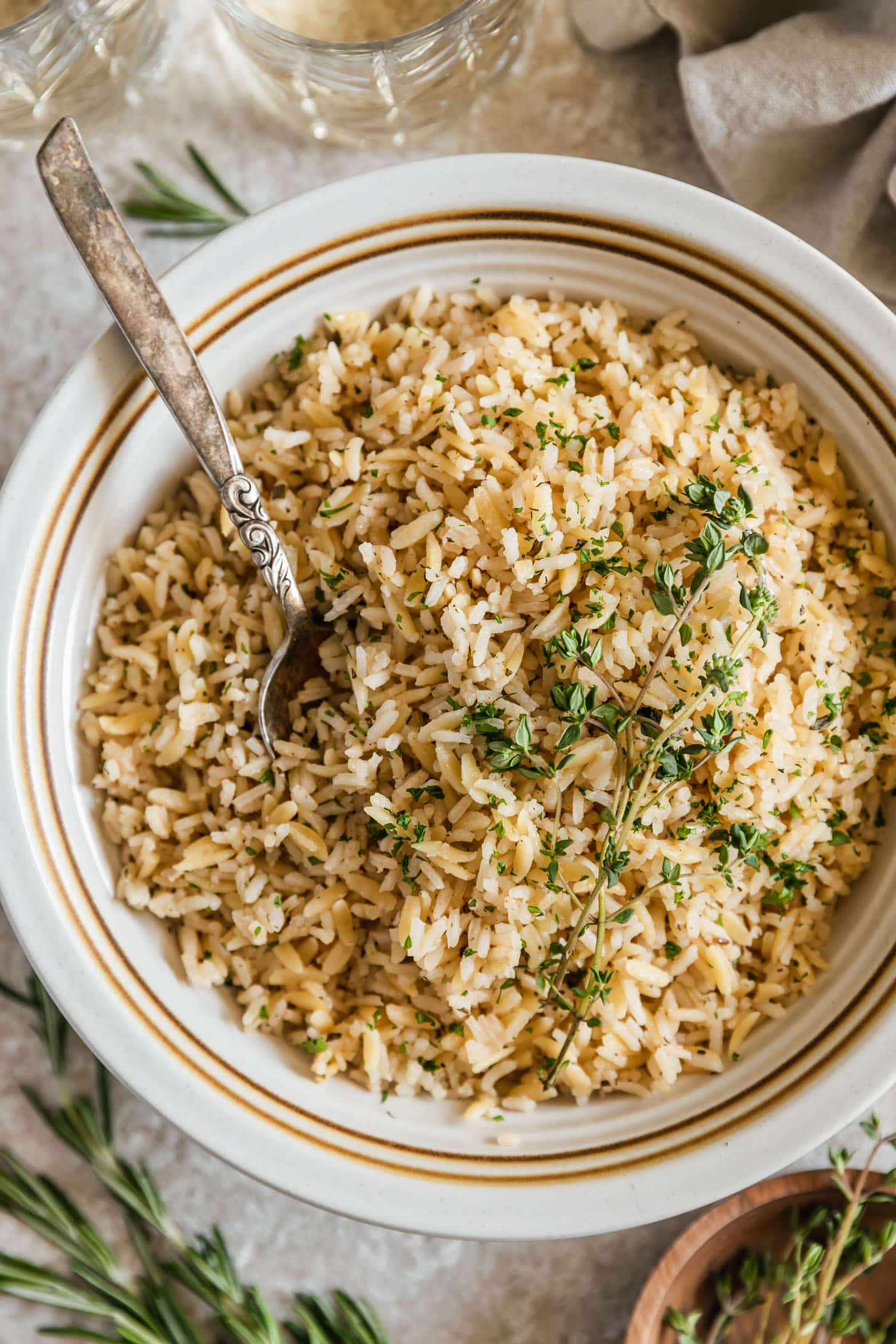 A white bowl of rice pilaf with orzo with a spoon on a beige counter next to glasses of white wine, thyme, and rosemary.