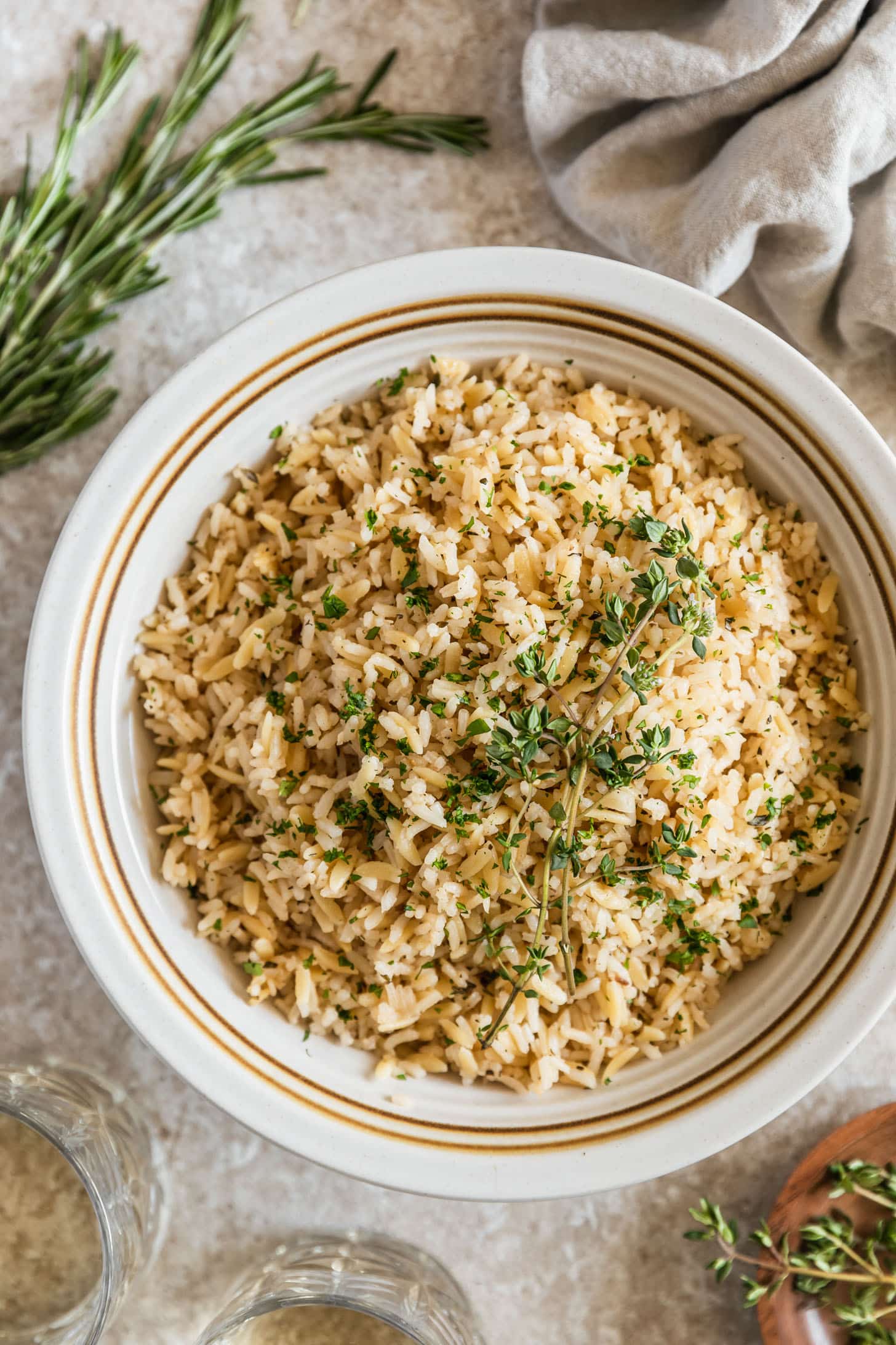 A white bowl of garlic herb rice pilaf with orzo on a beige counter next to thyme, rosemary, glasses of white wine, and a beige linen.