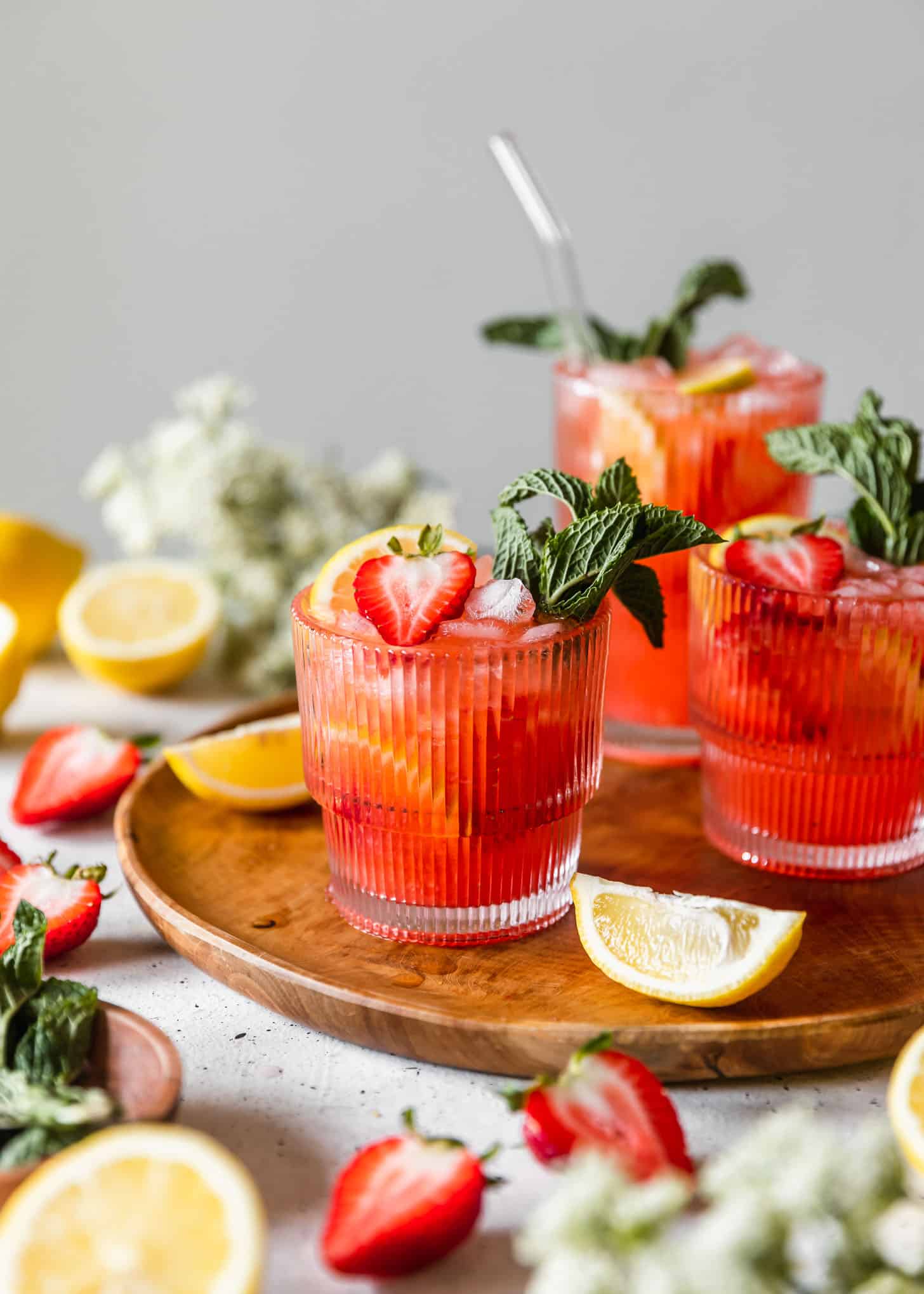 Three glasses of spiked strawberry lemonade on a wood tray next to white flowers, strawberries, and lemon halves on a white counter.